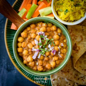 A green bowl filled with chole and topped with onions and cilantro; served with salad and parathas in the background.