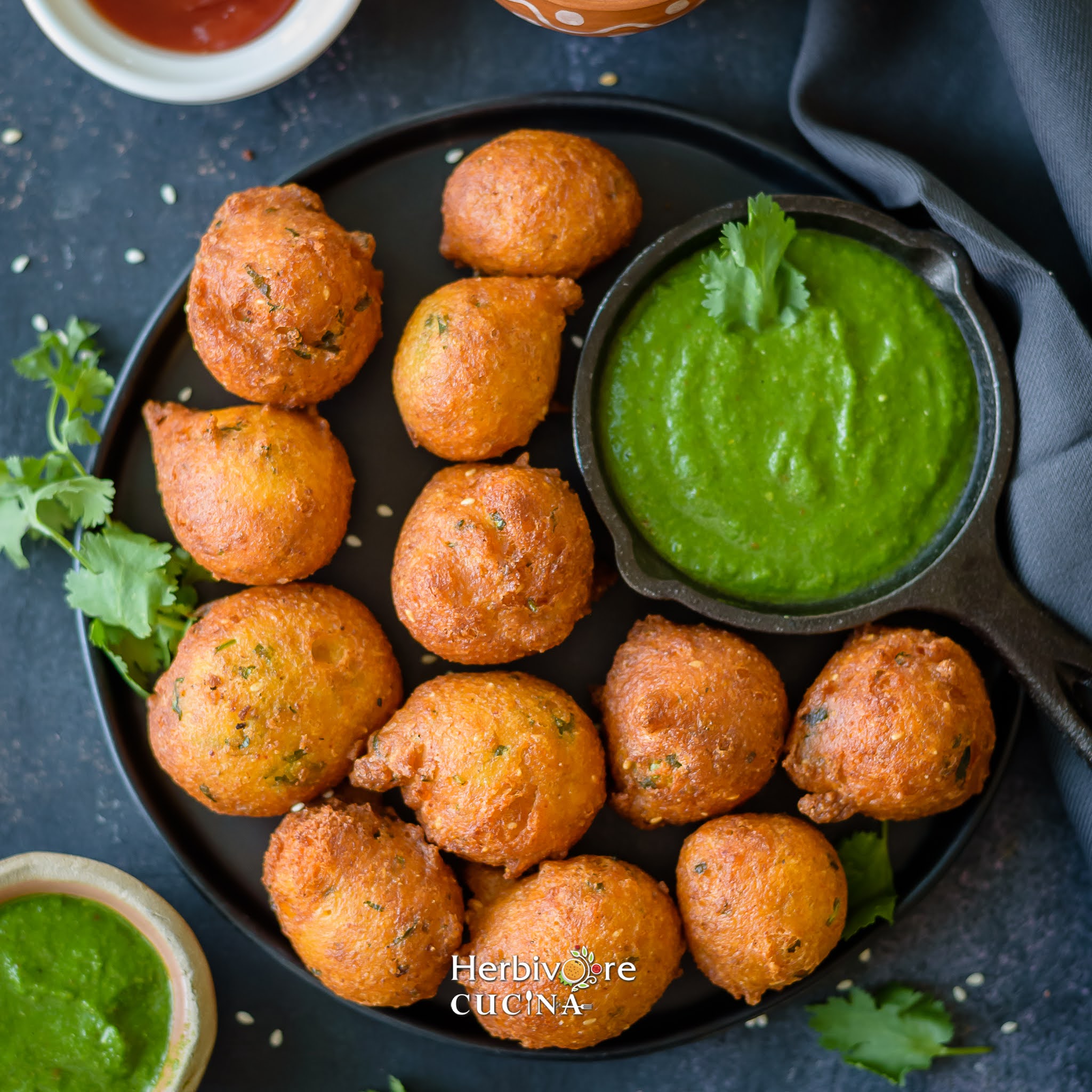 A black plate filled with crispy jowar and wheat vada along with green cilantro chutney with other chutneys in the background on a dark backdrop.