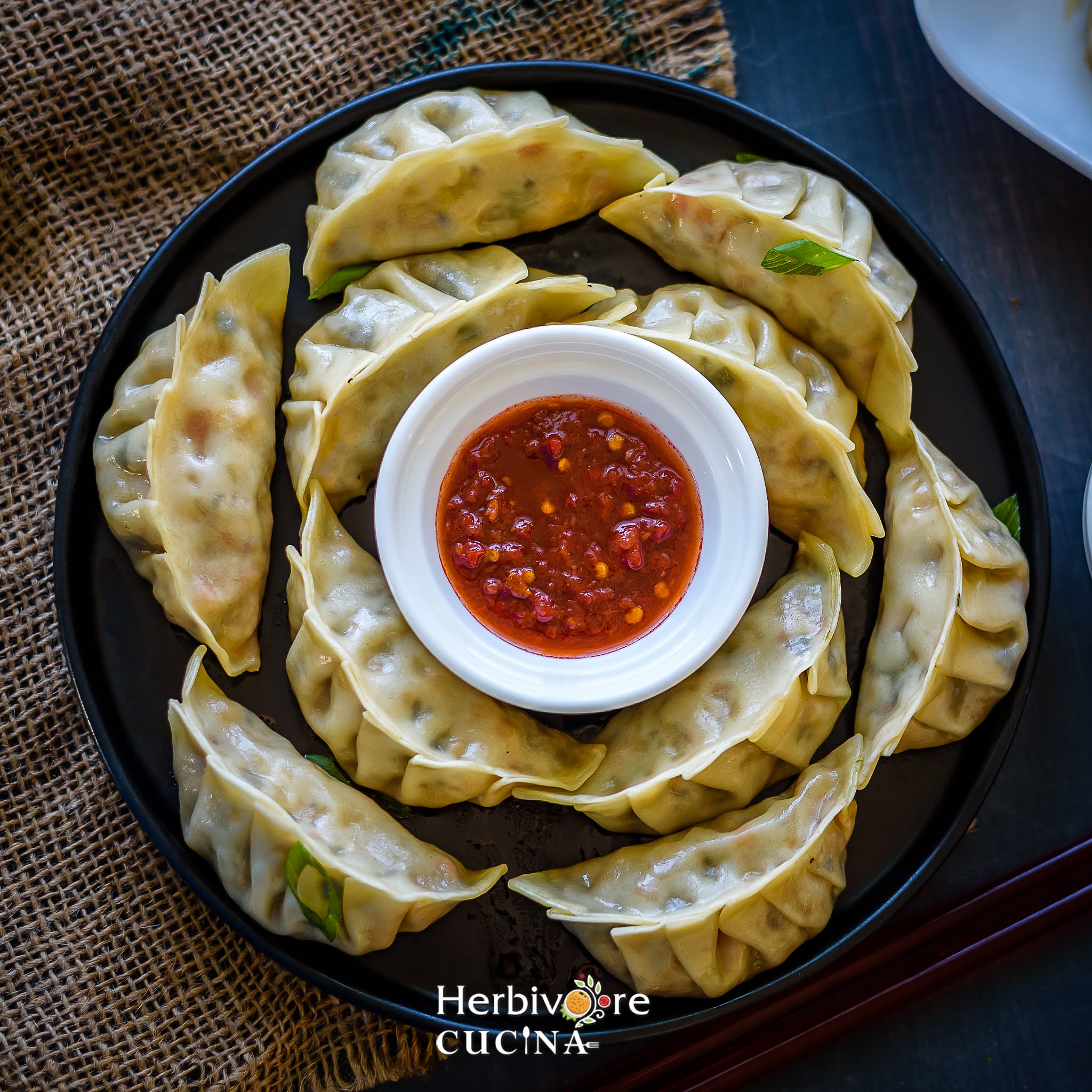 A black plate with pan fried dumplings and a bowl of hot sauce on a brown and black surface. 