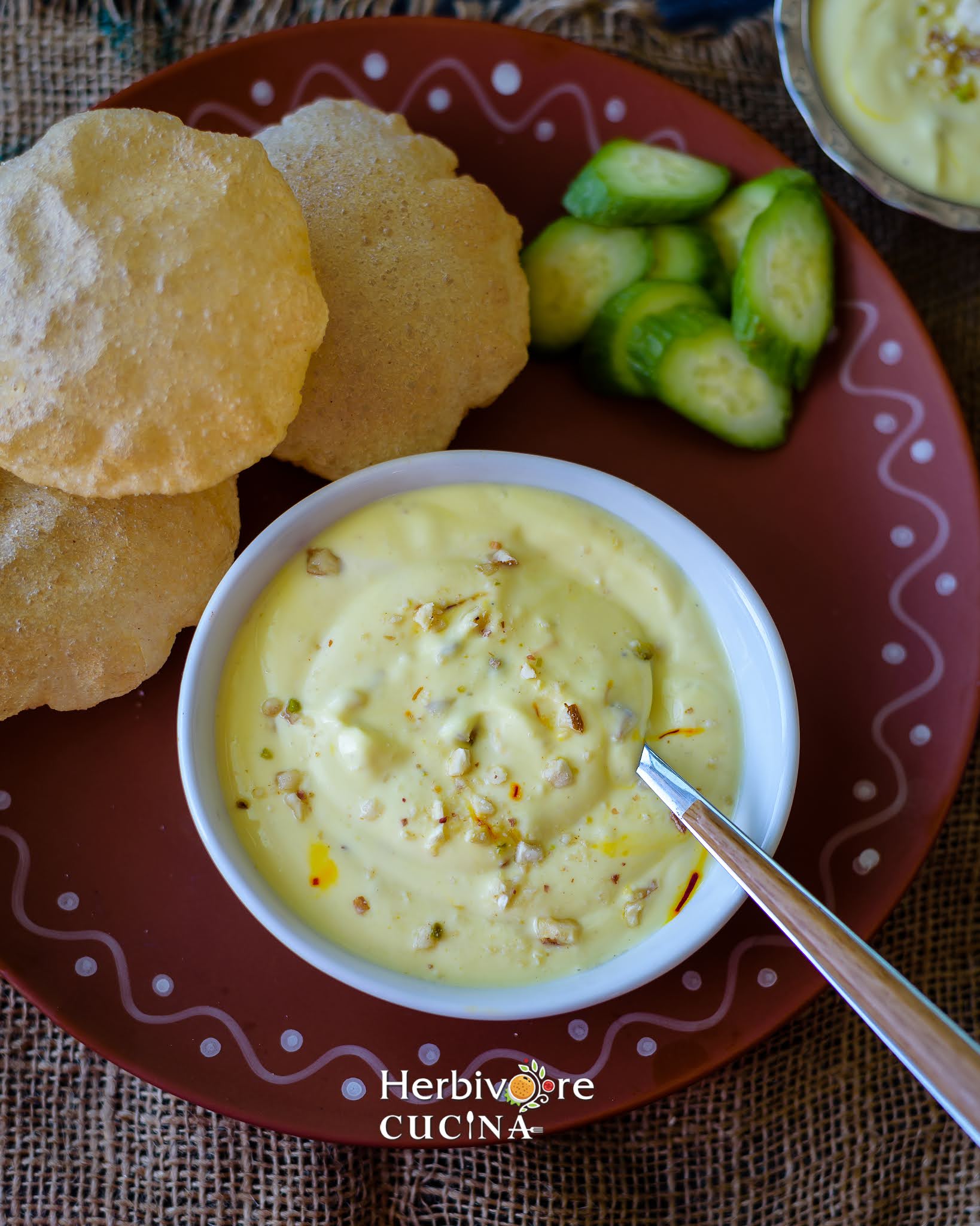 Kesar Shrikhand in a bowl with puri and cucumber in the background. 