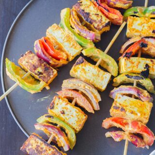 A black plate with tofu tikka arranged on three skewers and served with cilantro chutney on the side.