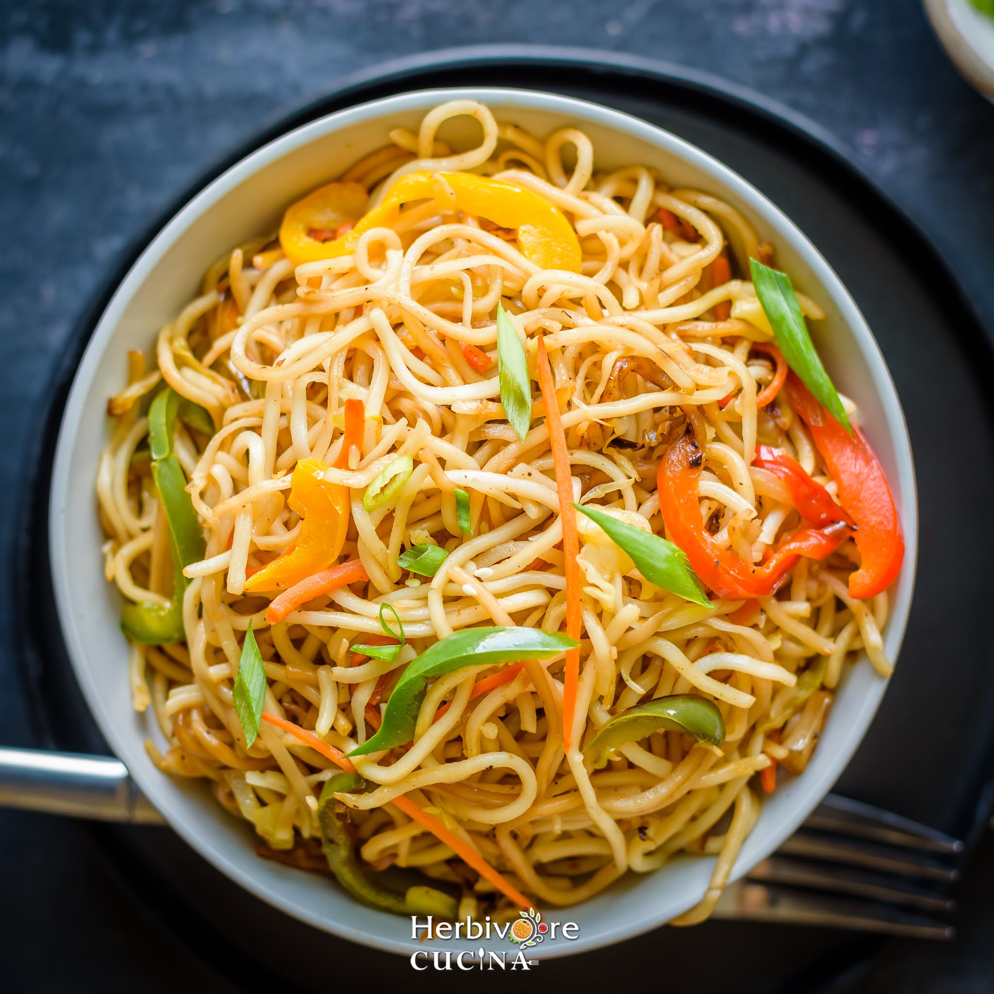 Top view of Hakka Noodles, a mix of noodles and vegetables with a fork on the side on a black background.