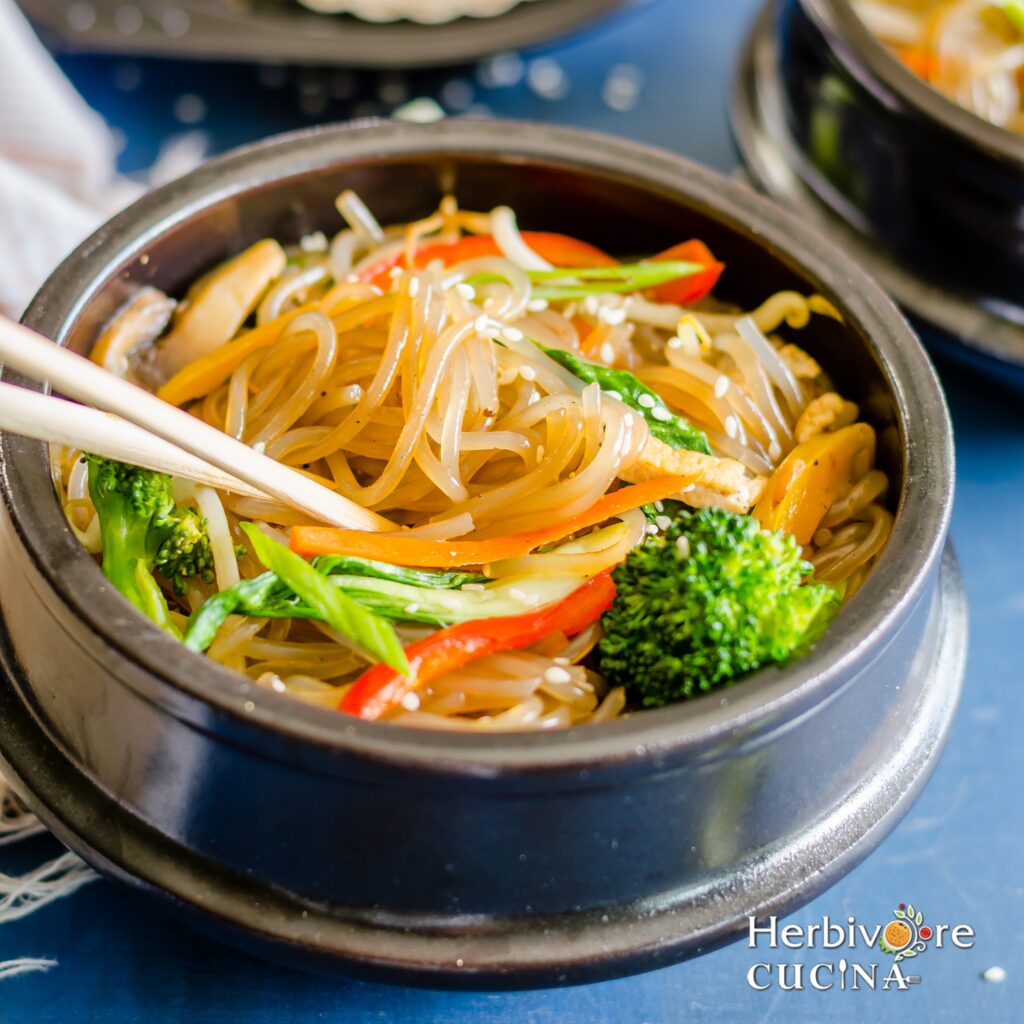 A black bowl filled with japchae; a sweet potato noodle stir-fry with vegetables and simple seasonings.