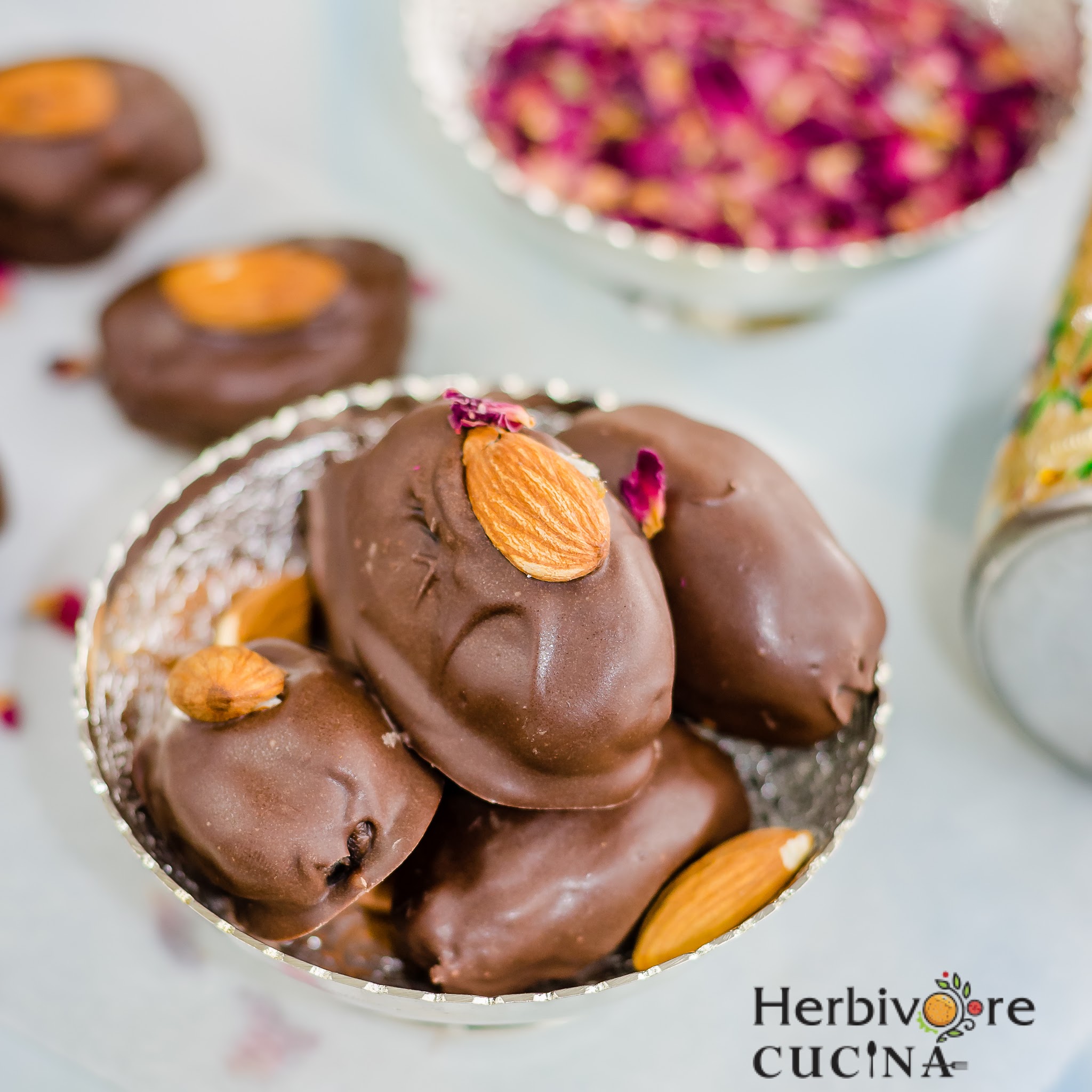 A silver bowl filled with chocolate covered dates topped with almonds and served with some rose petals on the side.