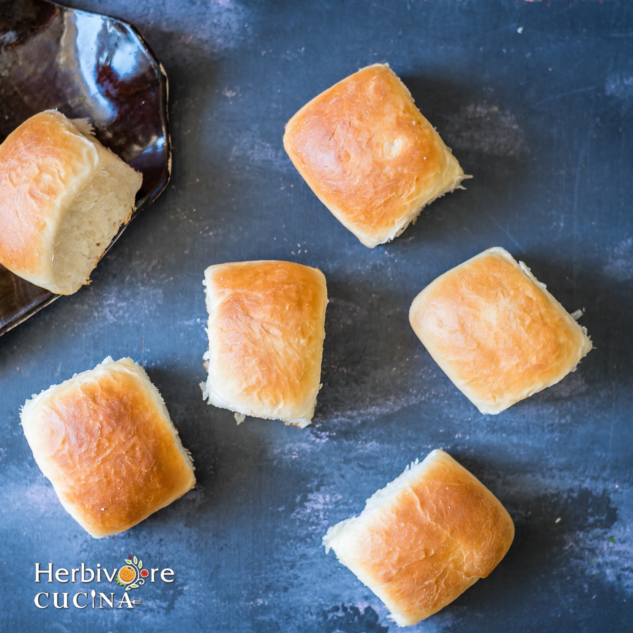 A dark surface with some pav breads on it and a tray with a bun on the side. 