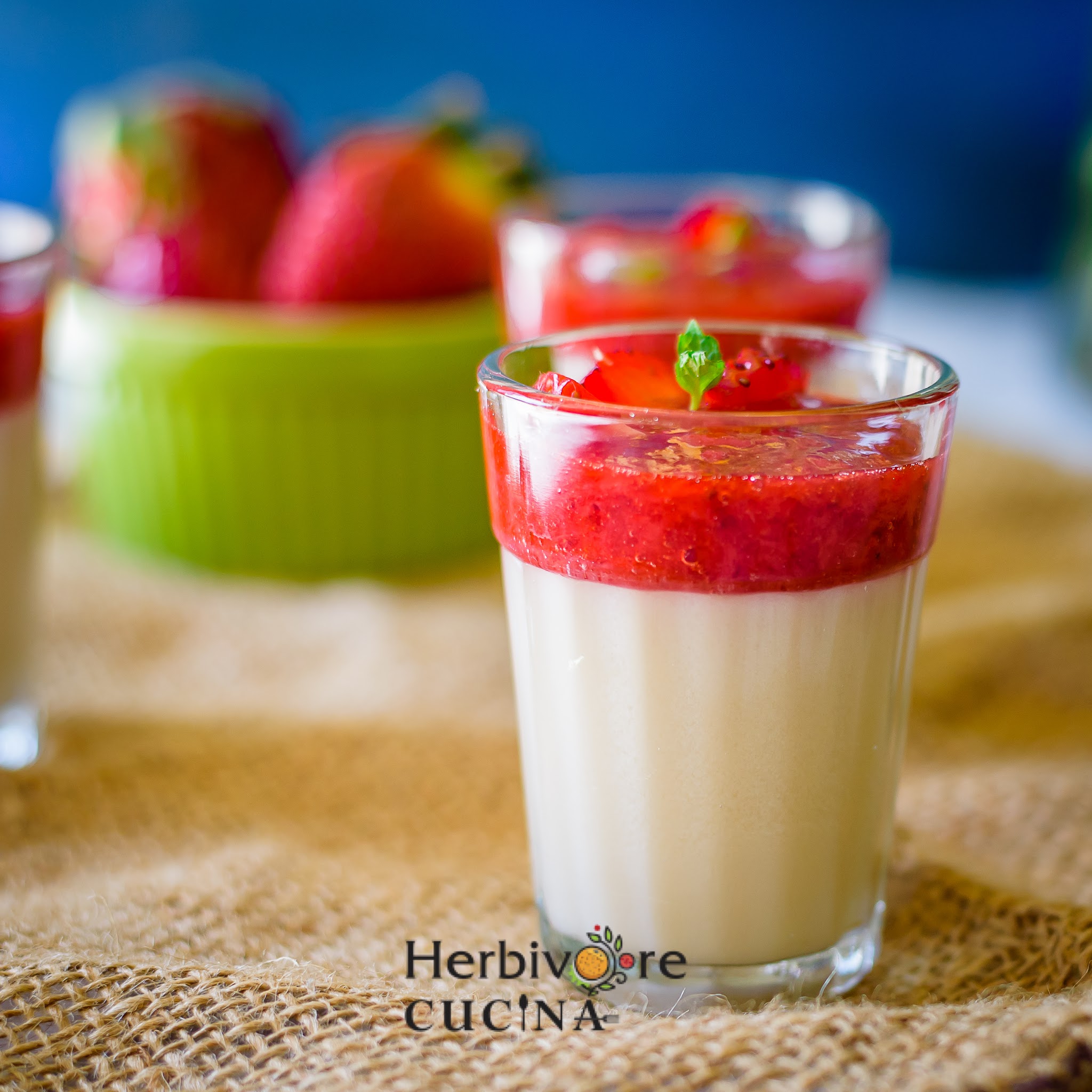 A glass with vanilla and strawberry panna cotta topped with strawberry jelly with more strawberries in the background. 