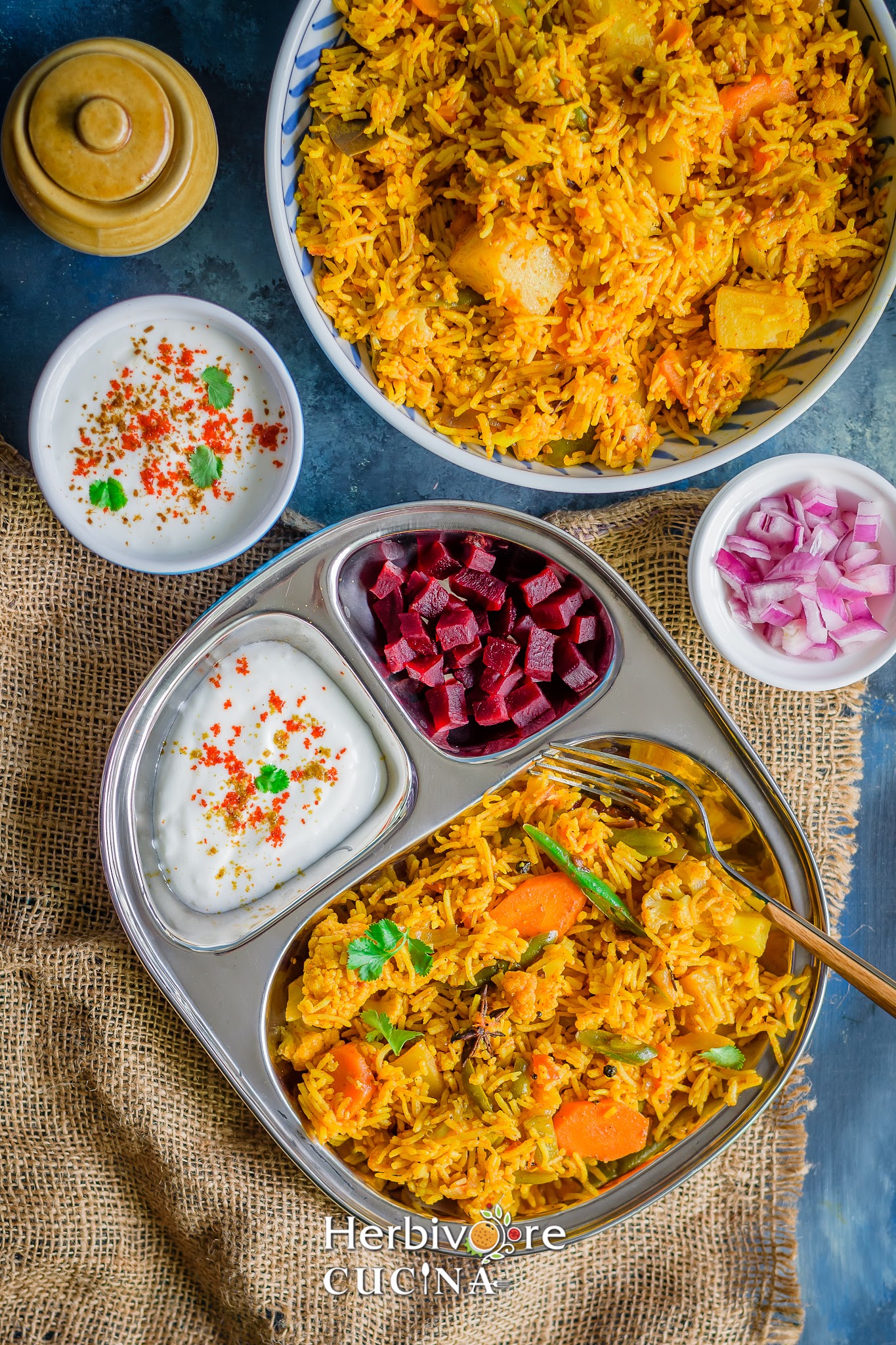 A steel plate with instant pot biryani, seasoned yogurt and chopped beetroots with more sides placed on a black board. 