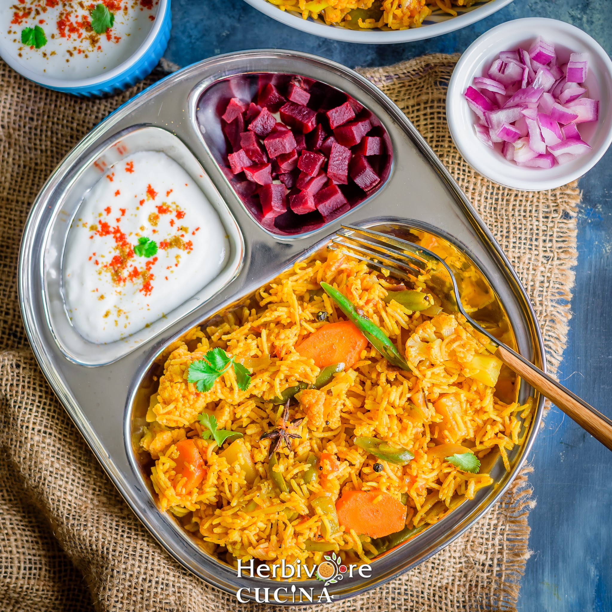 A steel plate with instant pot biryani, seasoned yogurt and chopped beetroots with more sides placed on a black board.