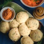 Overhead view of rava idli with tomato chutney, sambar and gun powder on a green brown plate.