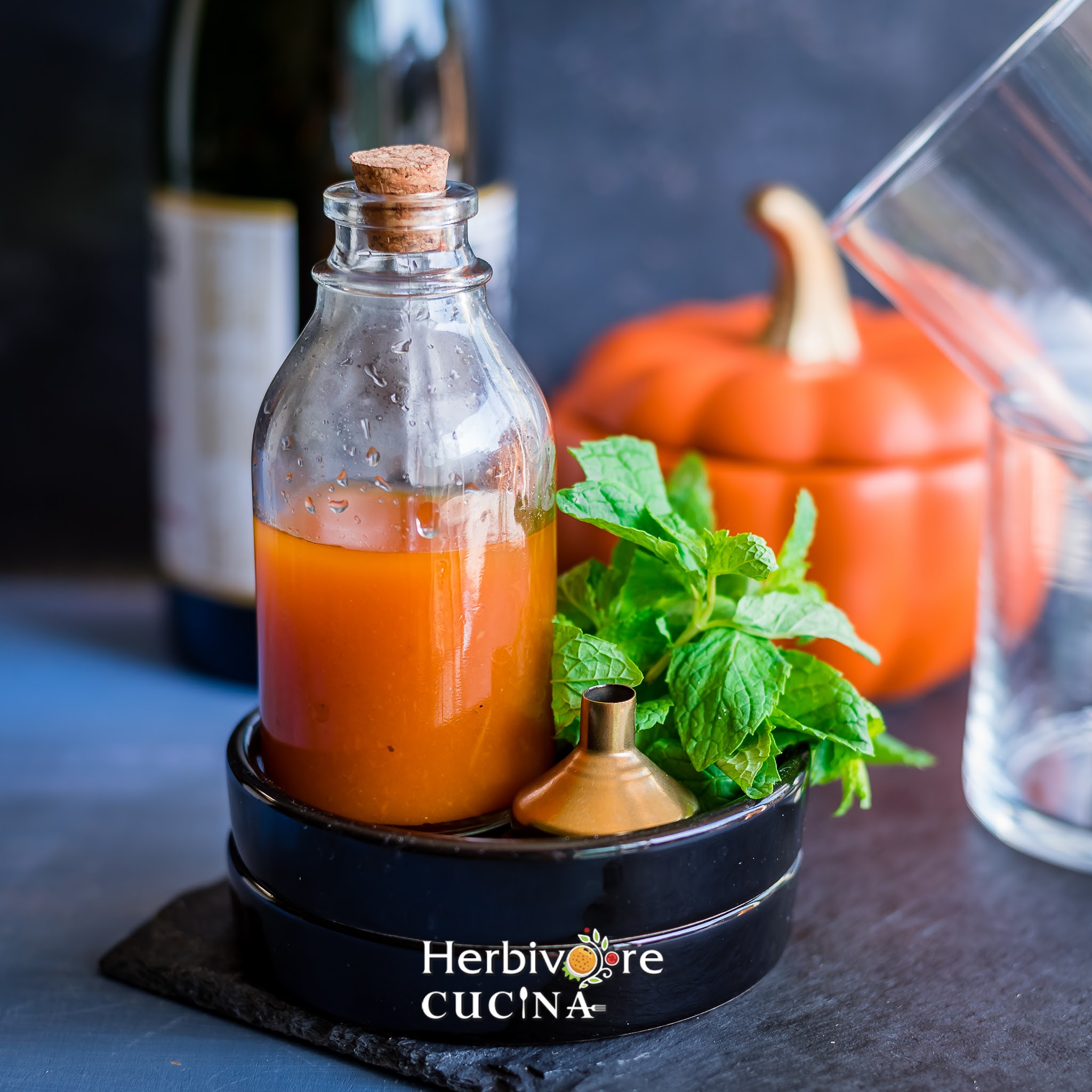Pumpkin simple syrup in a glass bottle with a funnel, mint leaves and glasses in the background.