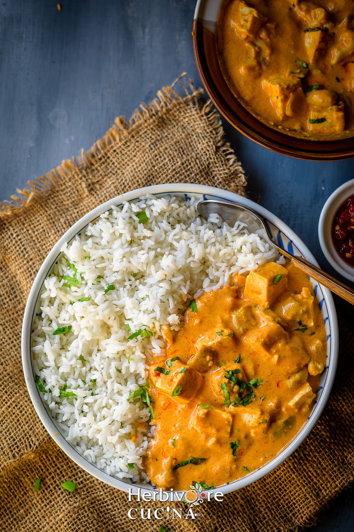 White plate with tofu peanut curry and steamed rice served with cilantro on top on a gray board with a brown surface under it. 