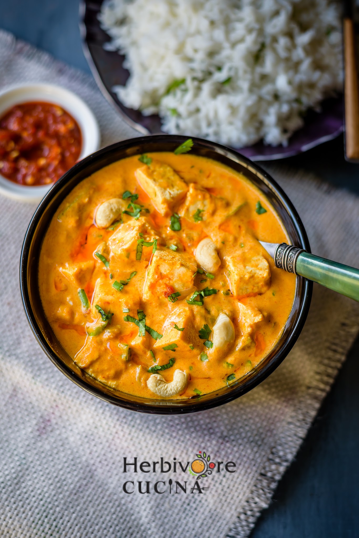 A black bowl with tofu and cashew curry topped with cilantro and with a fork by the side and served with hot sauce and steamed rice by it. 