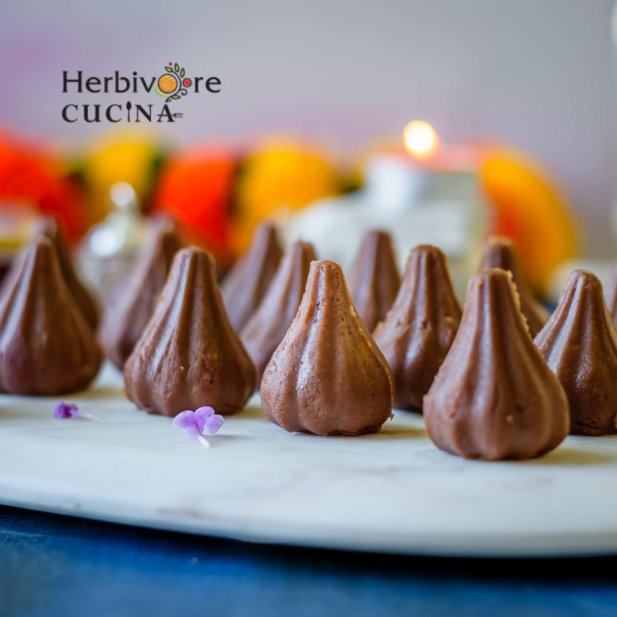 Chocolate modaks arranged on a marble platter with a candle and flowers in the background.