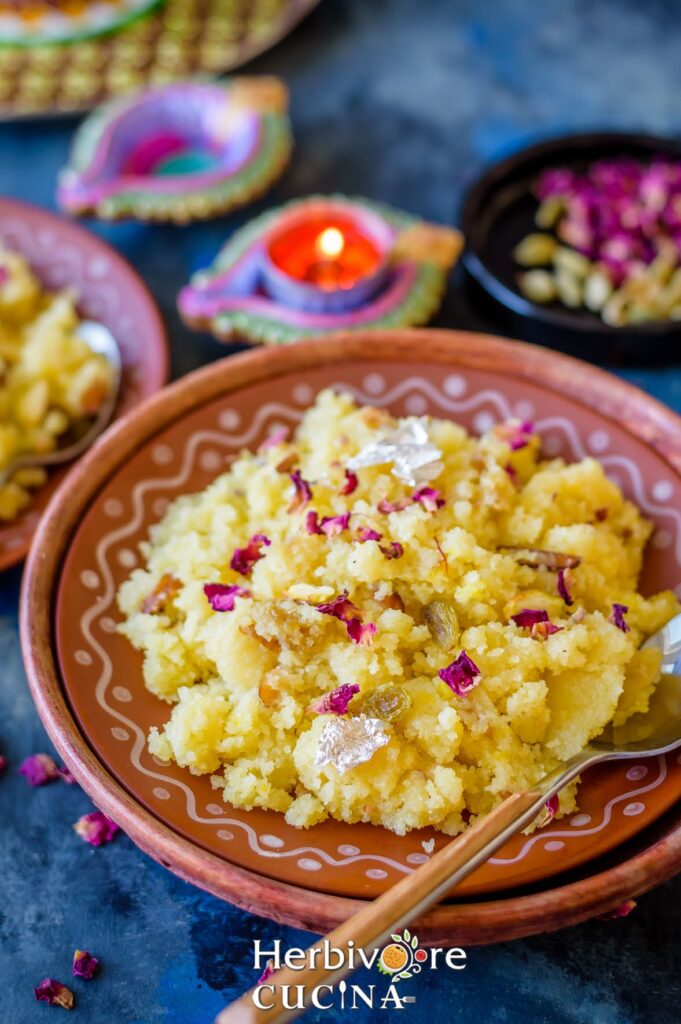 A brown plate with sooji halwa served topped with rose petals on top; with diyas on the background.