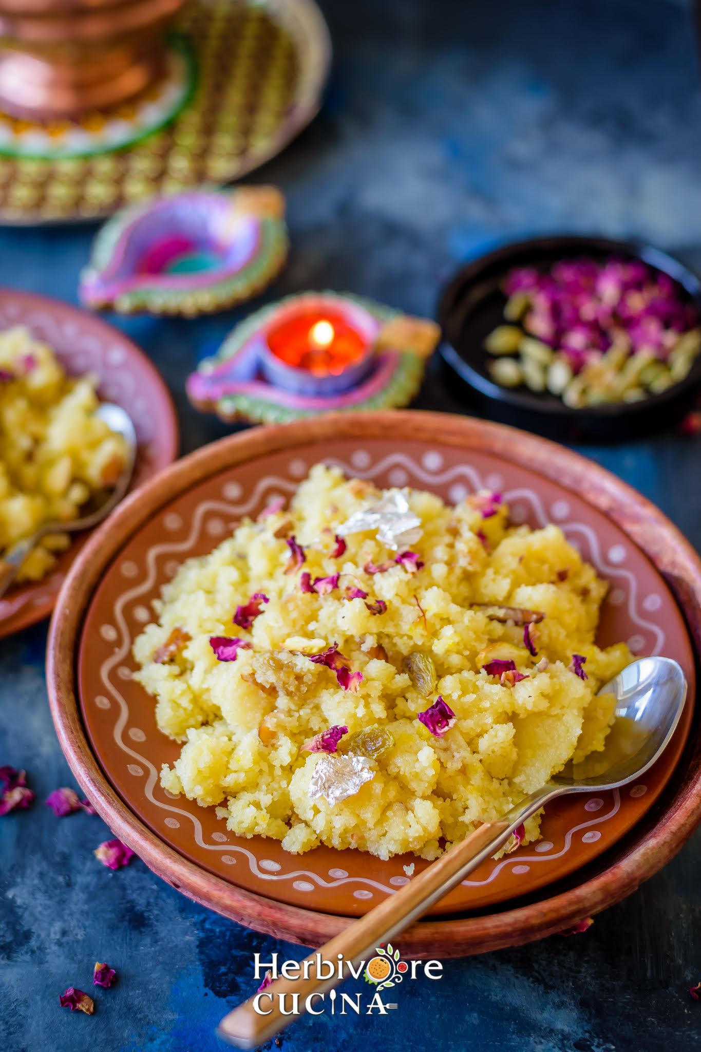 A brown plate with sooji halwa served topped with rose petals and saffron with diya in the background. 