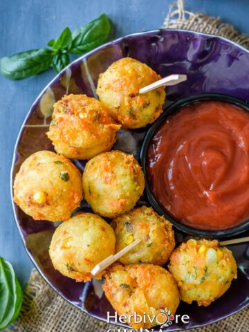 A violet plate filled with baked cheese balls with a small bowl of ketchup by the side on a gray board.