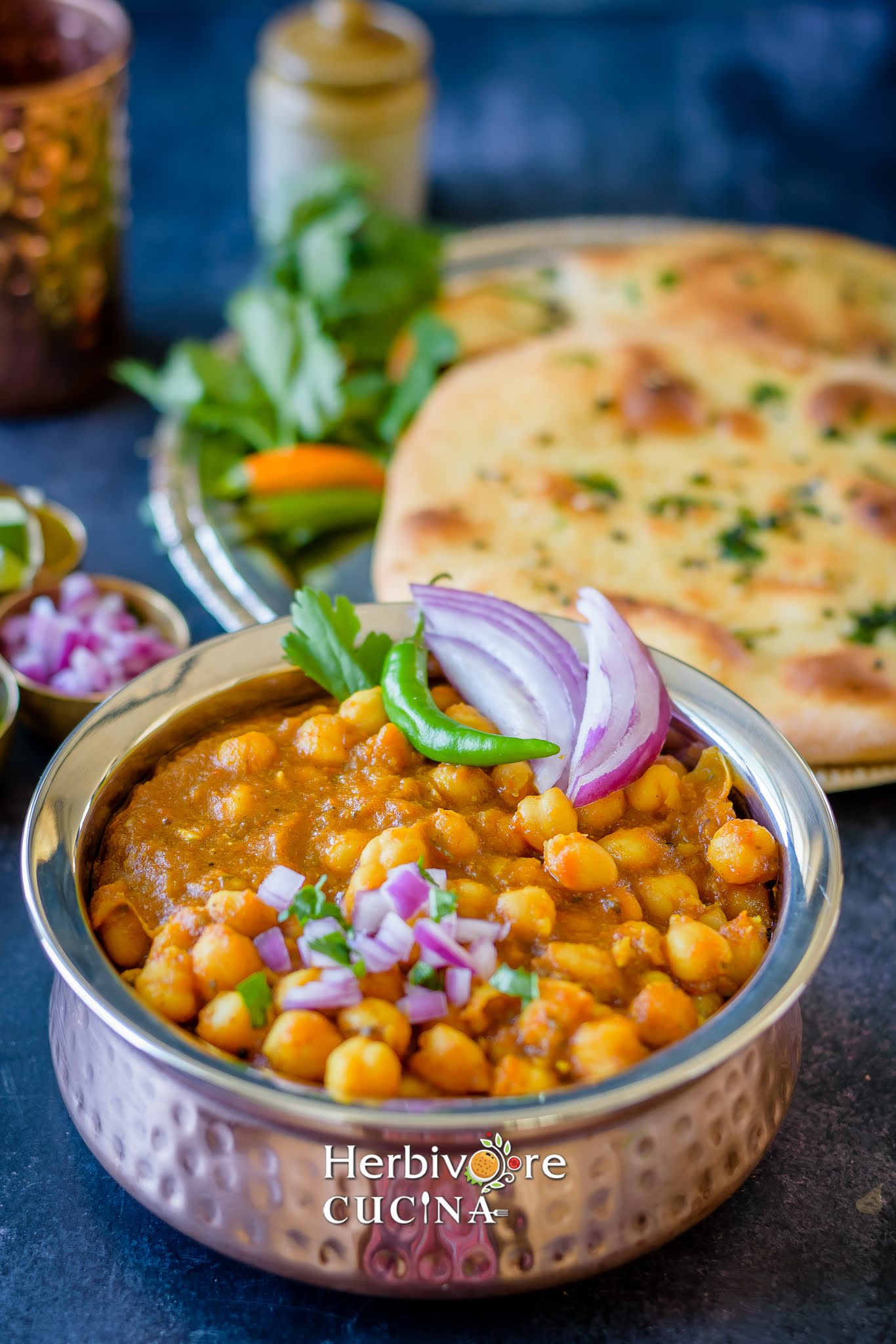 A handi filled with chole and topped with onions and chili with kulcha in the background. 