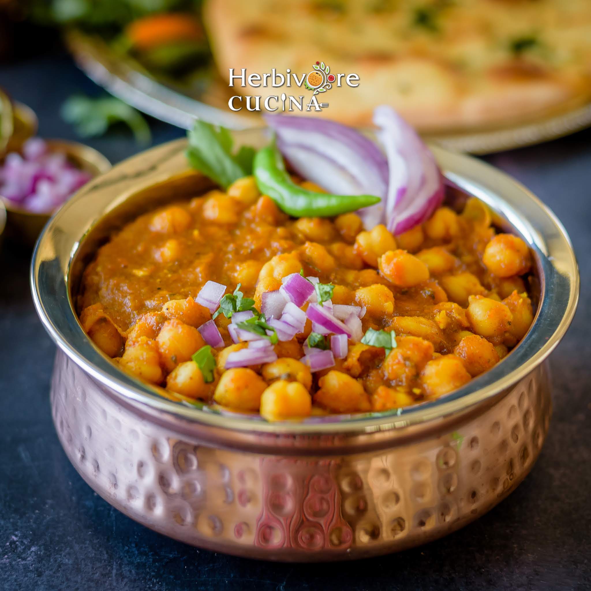 A copper vessel filled with chole and topped with diced onions, green chili and cilantro; with kulcha on the side.