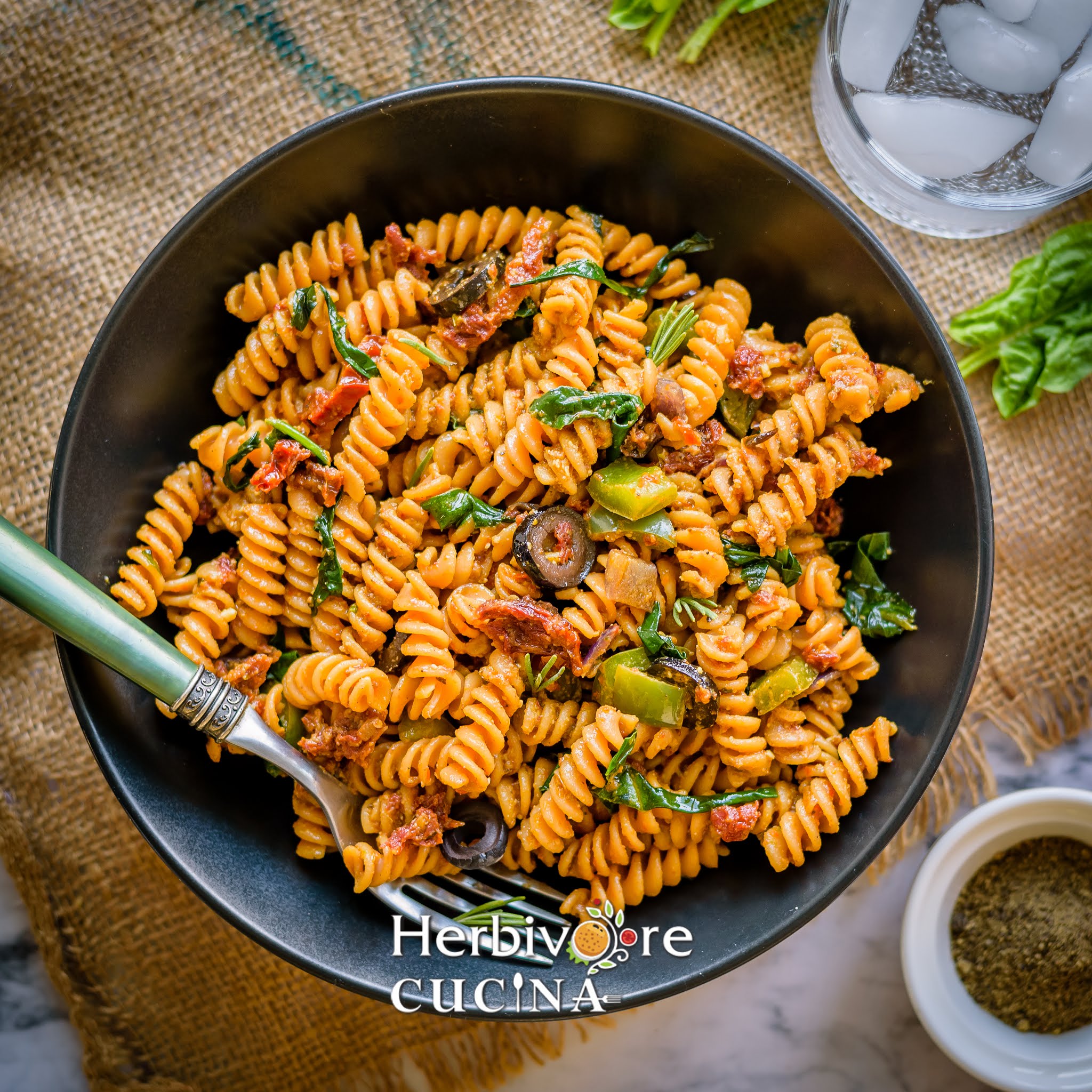 A black bowl with red lentil pasta tossed in sun-dried tomato pesto with spices on the side.