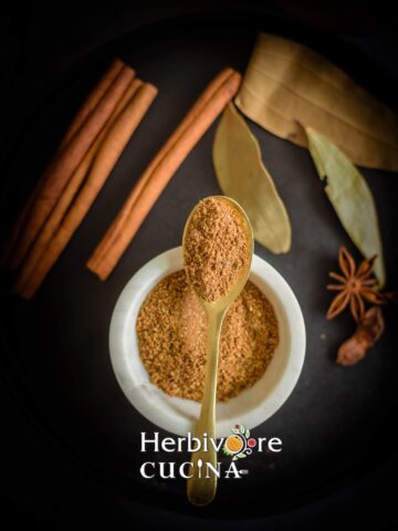 A white bowl with garam masala in it with a golden spoon above it with some masala on a black background.