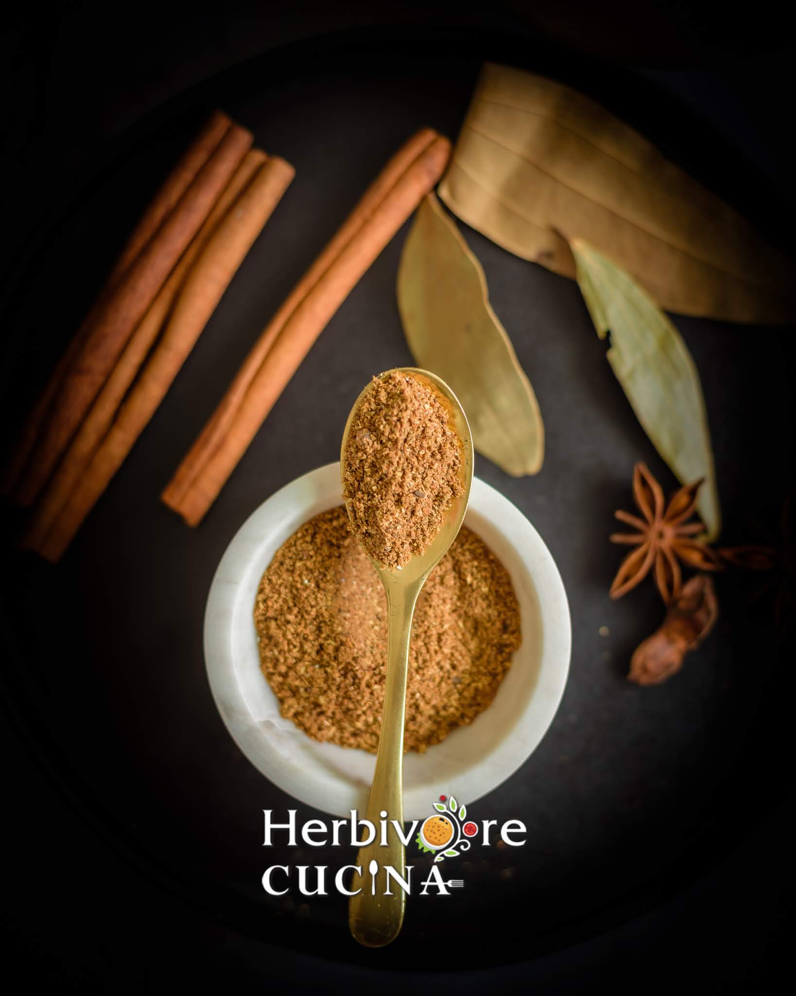 A white bowl with home-made garam masala with a golden spoon filled with garam masala above it on a black background. 