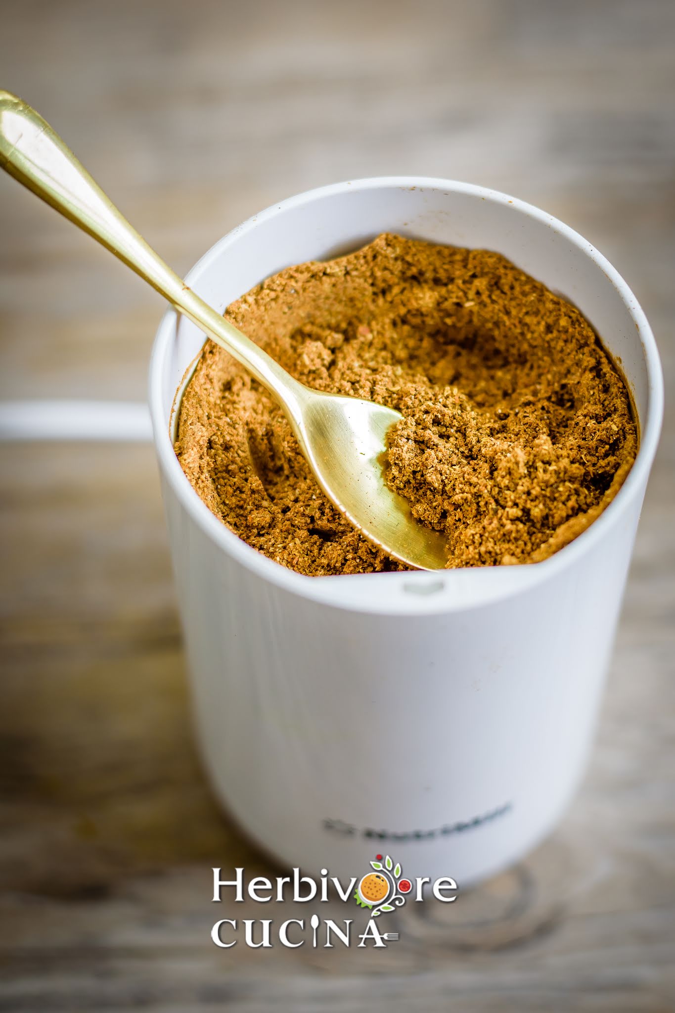 A spice mixer filled with garam masala and a golden spoon in it on a brown background. 