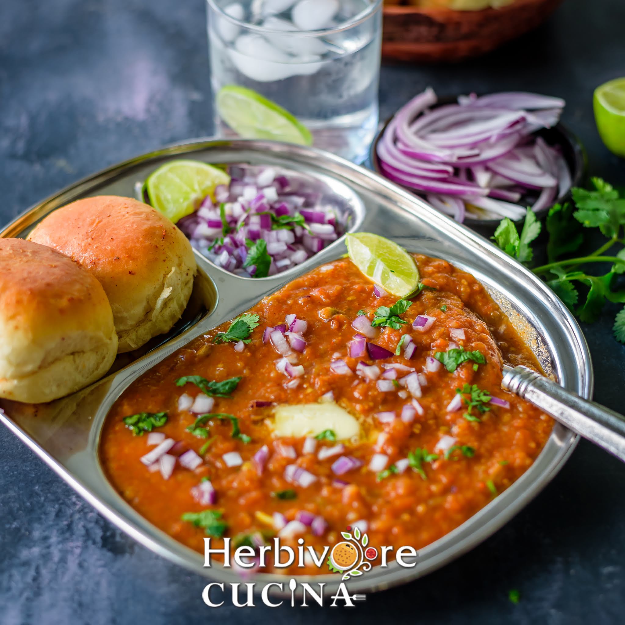 A steel plate with pav bhaji served with chopped onions and cilantro with butter on the bhaji.