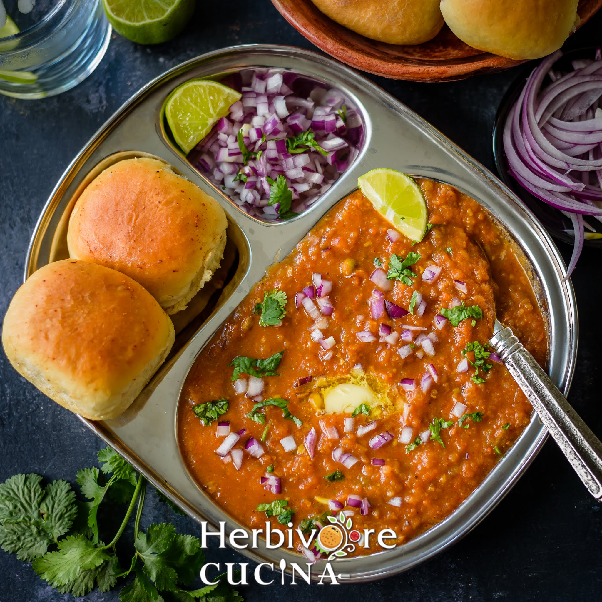 A steel plate filled with bhaji and served with pav, onions and lemon on a black board. 