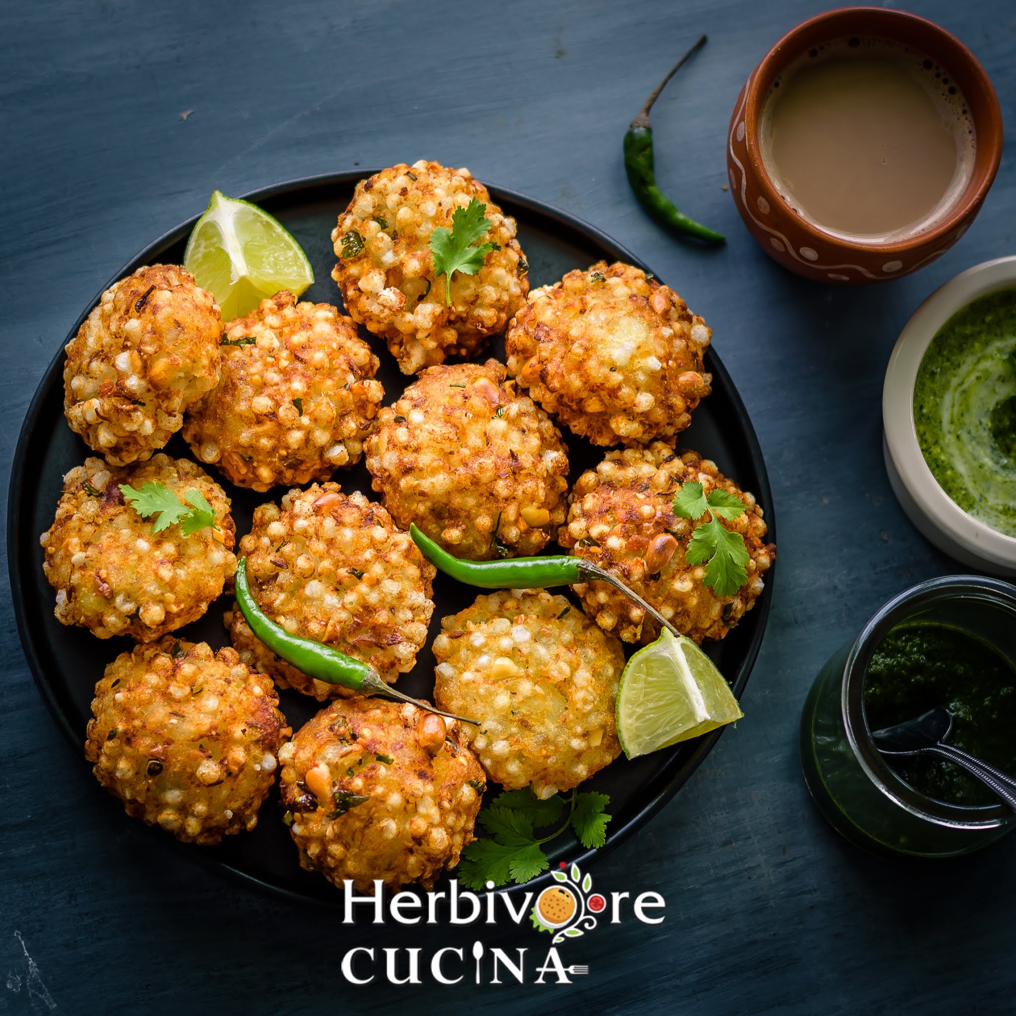 Deep fried Sabudana Vada arranged in a black plate with some lemon slices and green chili around it with masala chai and chutney around it. 