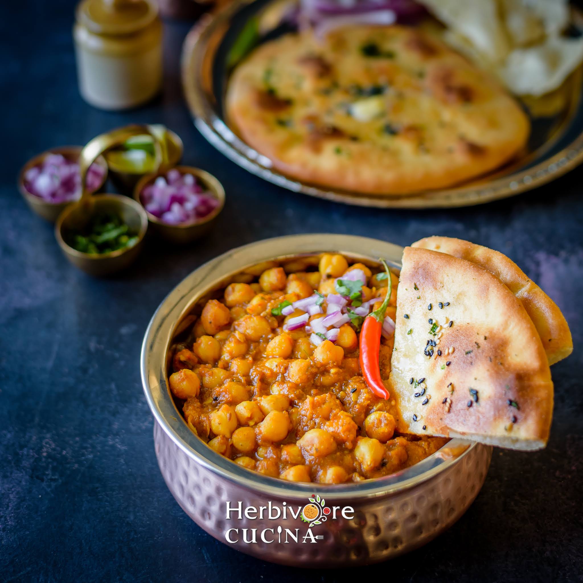 A bowl of chole with a piece of kulcha on the side with more kulcha in the background. 