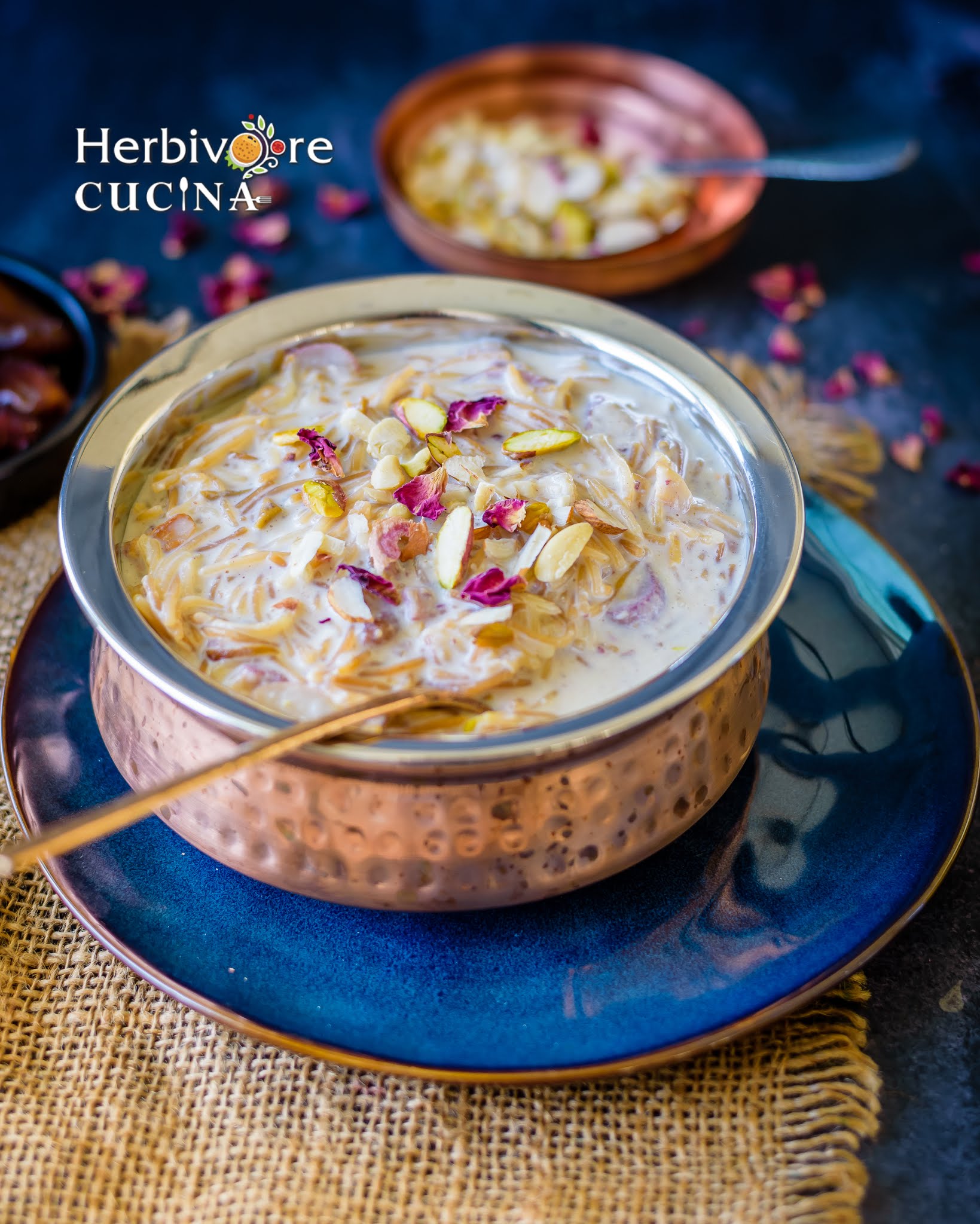 A copper container with sheer khurma topped with rose petals and nuts on the side.