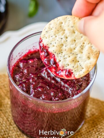 Glass container filled with mixed berry jam and a crisp dipping in it.