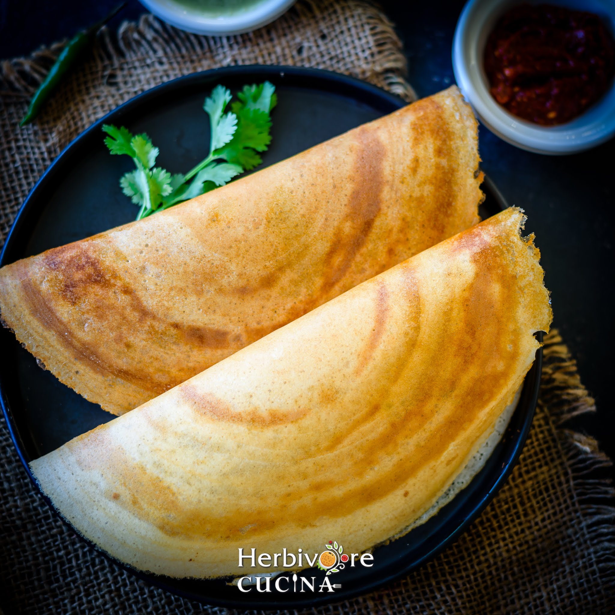 A black platter with two butter dosas and some chutneys on the side.