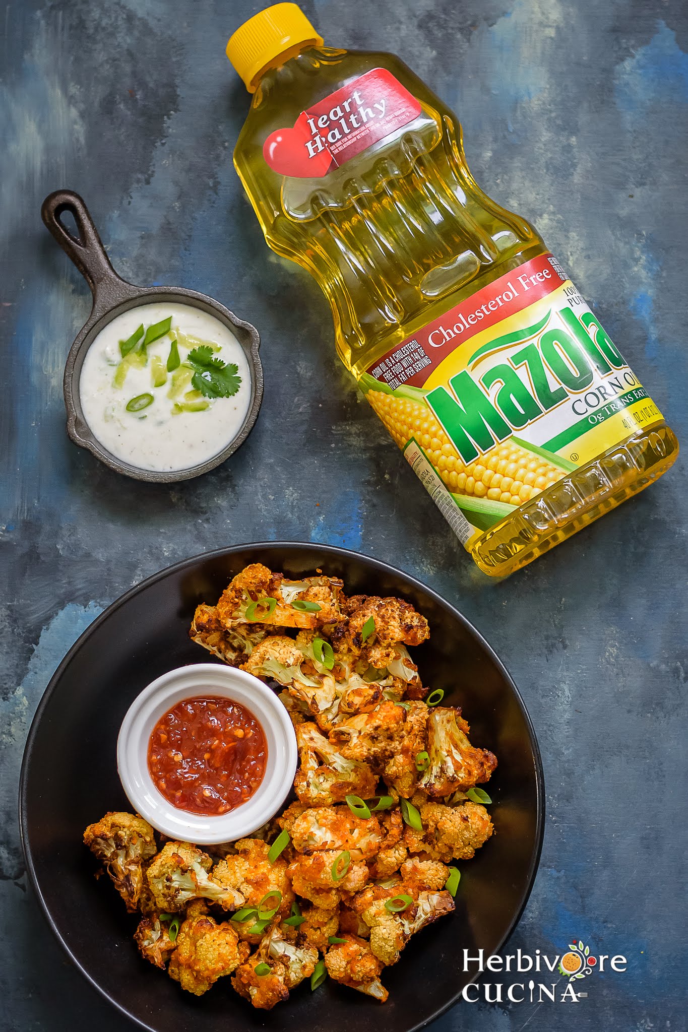 A black bowl filled with air fryer cauliflower bites and some sweet chili sauce with a yogurt dip in a black bowl and a bottle of corn oil on the side. 