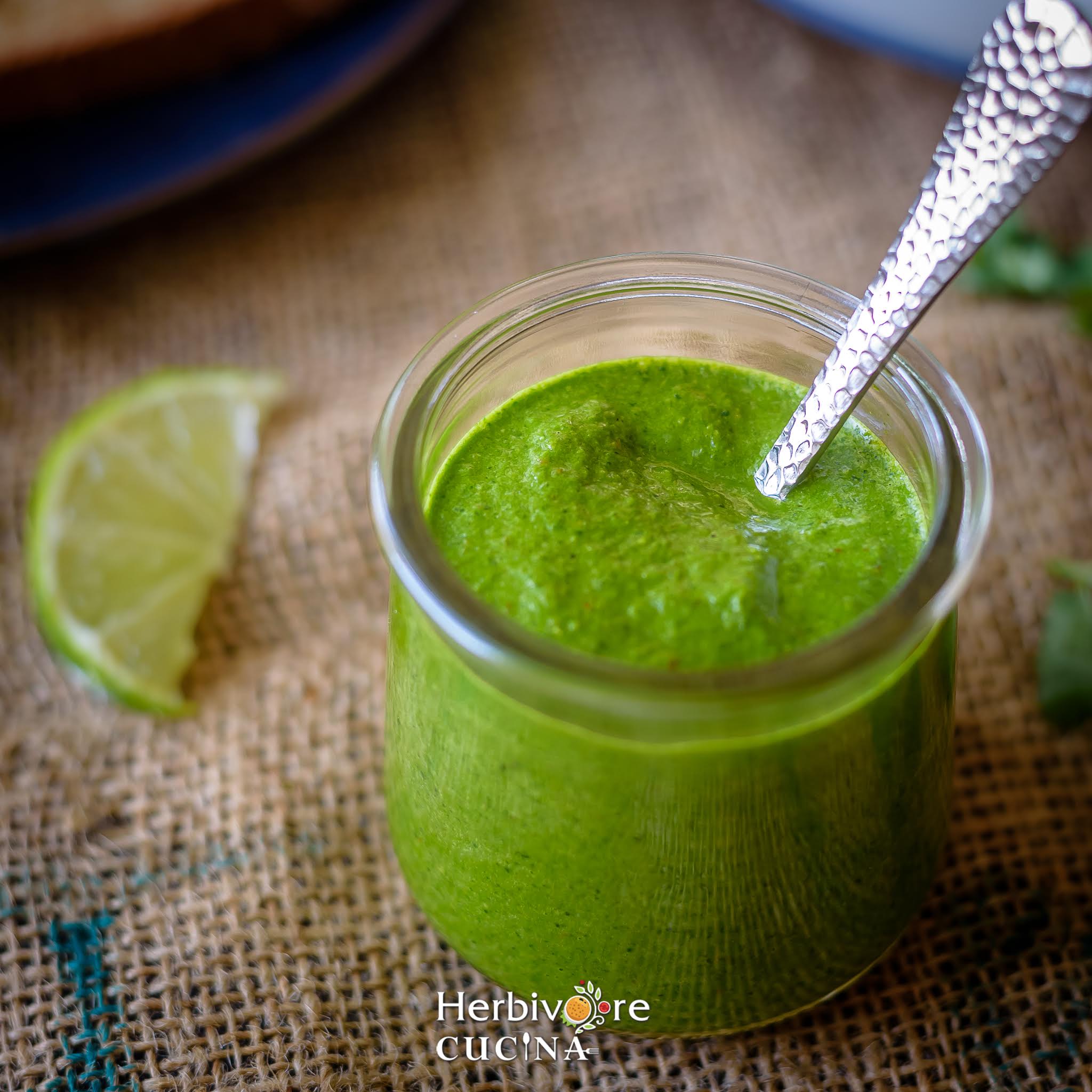 A glass bottle with cilantro chutney and a steel spoon in it on a brown background with lemon slice beside it. 