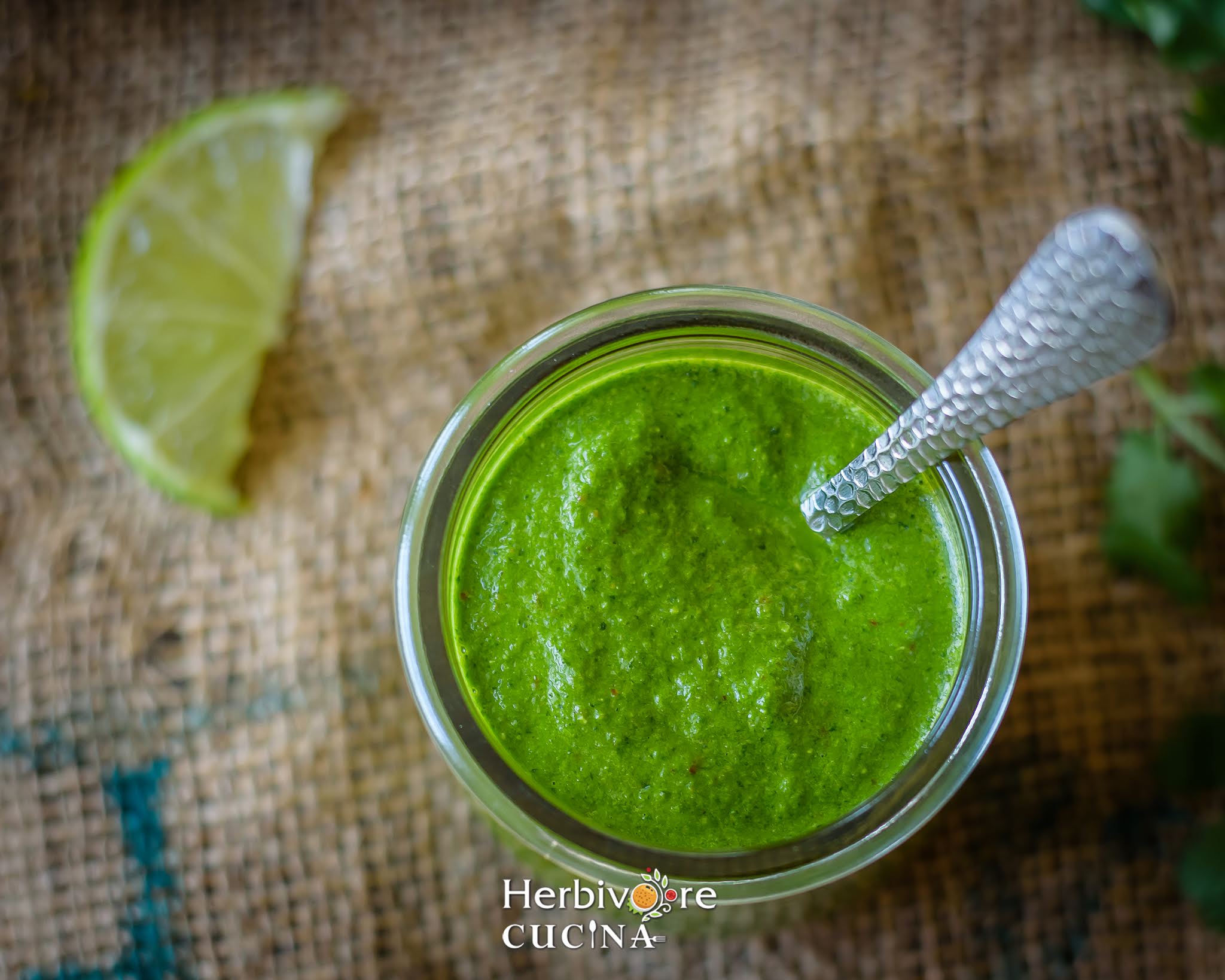 A glass jar with cilantro chutney on a gunny bag with lime slice beside it and a spoon in the jar. 