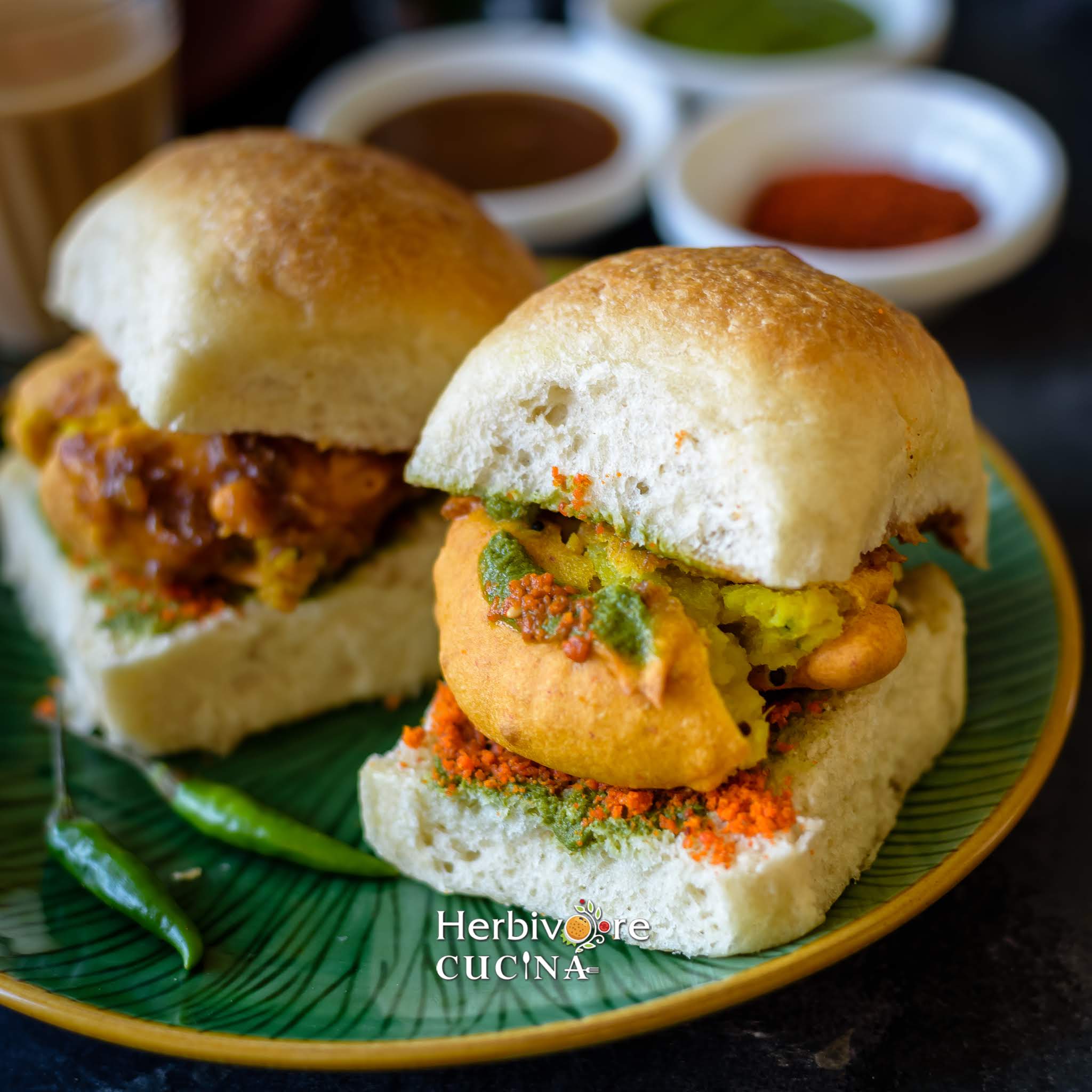 Vada Pav served in a green plate with more vada pav in the background and chutneys served in bowls placed behind it.