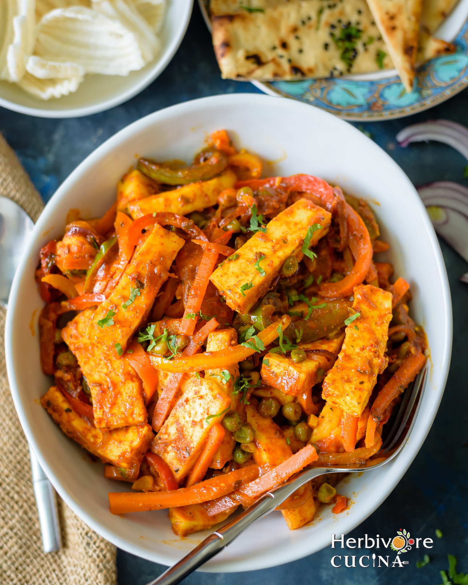 Paneer Jalfrezi; a delicious mix of vegetables and paneer in a tomato based sauce served in a white bowl with naan and papad on a blue background.
