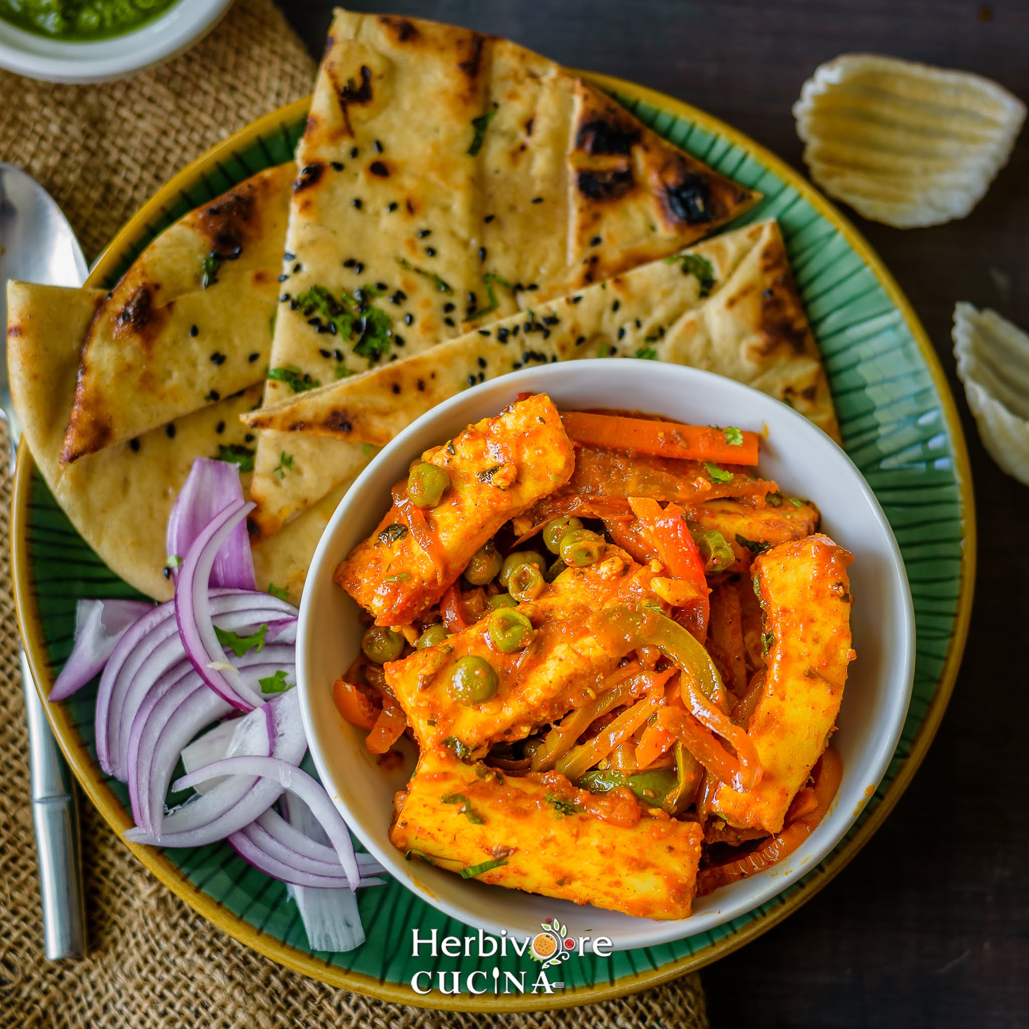A white bowl filled with paneer jalfrezi served with naan and sliced onions in a green plate with papad on the side. 
