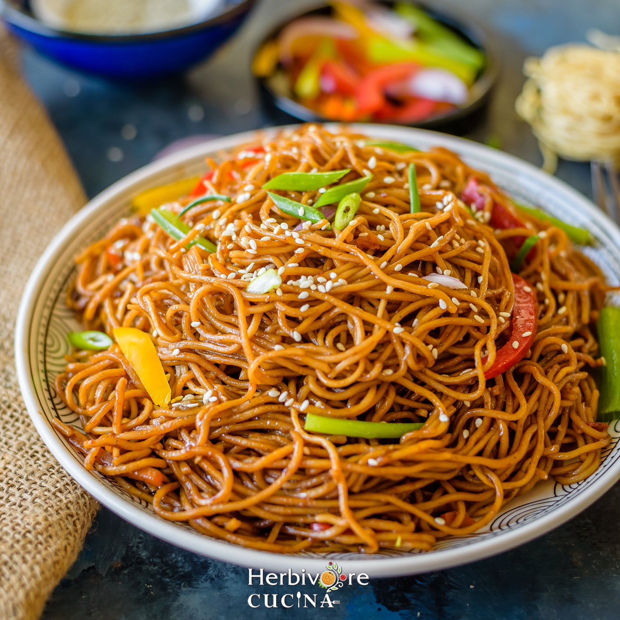 Teriyaki Noodles served in a white plate topped with sesame seeds and some chopped vegetables seen in the background. 