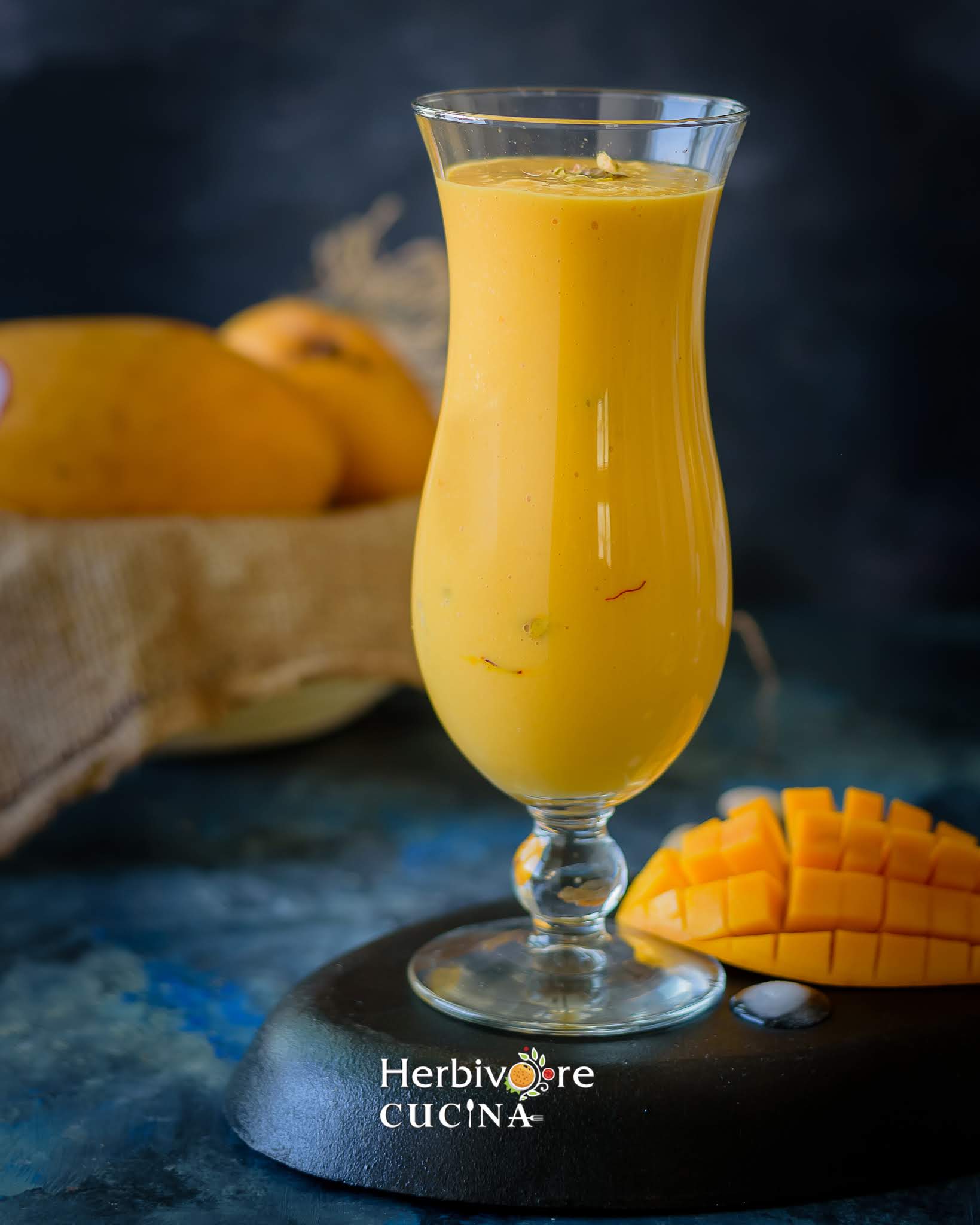 A tall glass of mango lassi with some whole and chopped mangos on the side against a dark background.