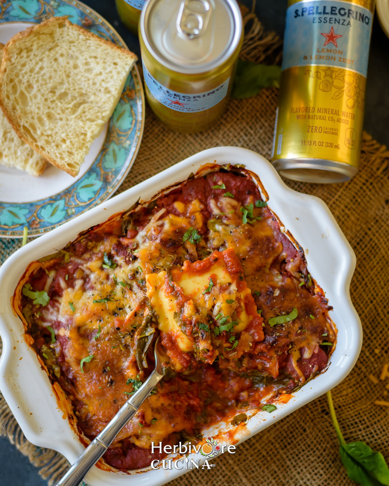 A white square bowl with ravioli lasagna picked with a spatula; with slices of bread and soda on the side.