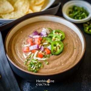 A black bowl with refried beans with colorful toppings with a bowl of chips on the side.