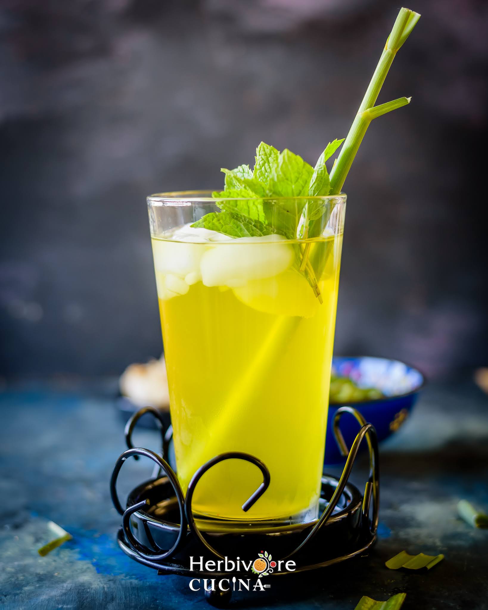 A glass full of lemongrass ginger cooler topped with ice and mint leaves against a blue background. 