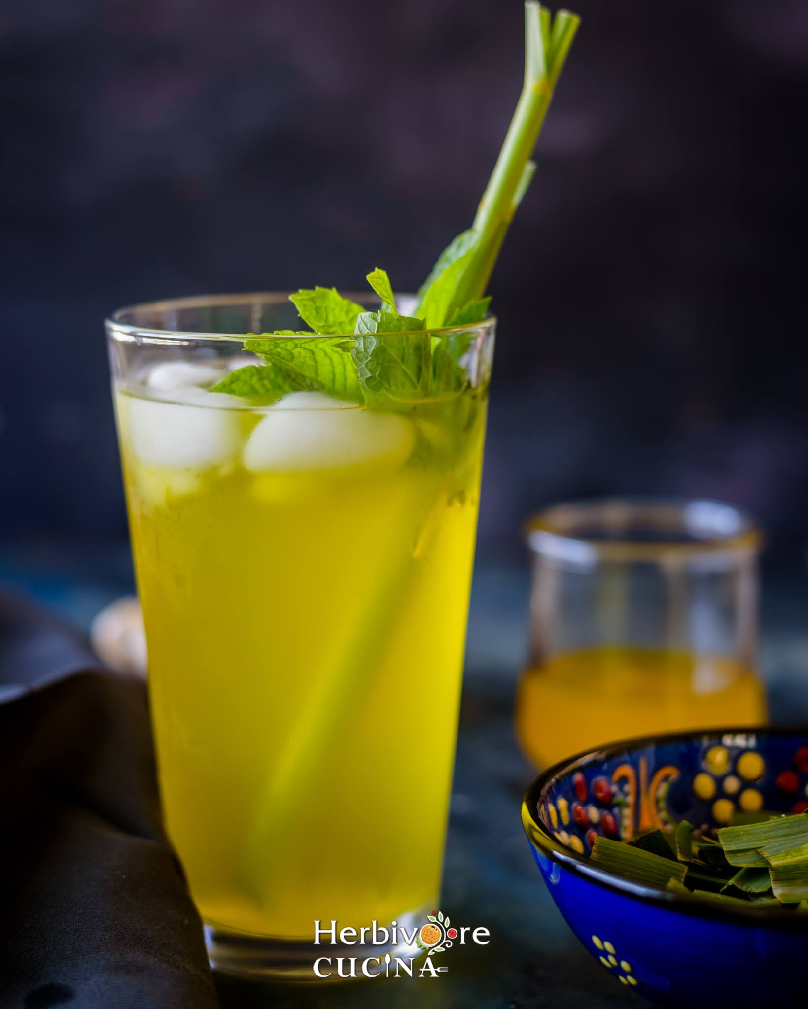 A tall glass filled with lemongrass ginger cooler topped with ice, mint leaves and lemongrass stalk against a dark background. 