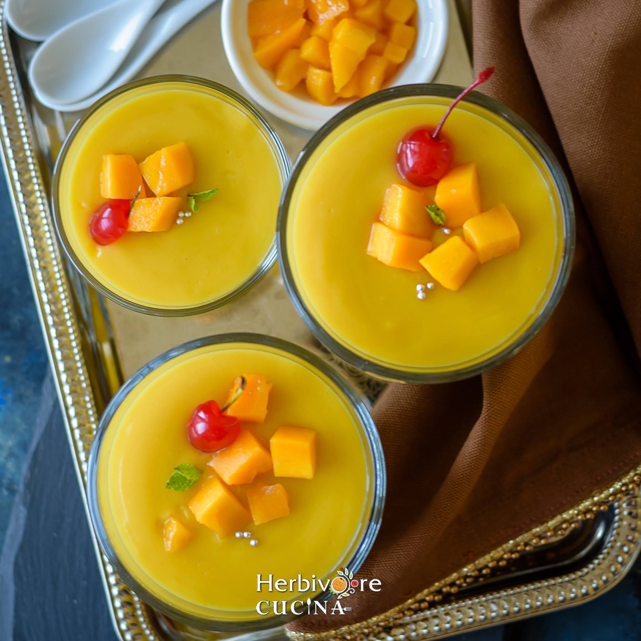 A tray with three servings of eggless mango custard pudding topped with mangos and cherries.