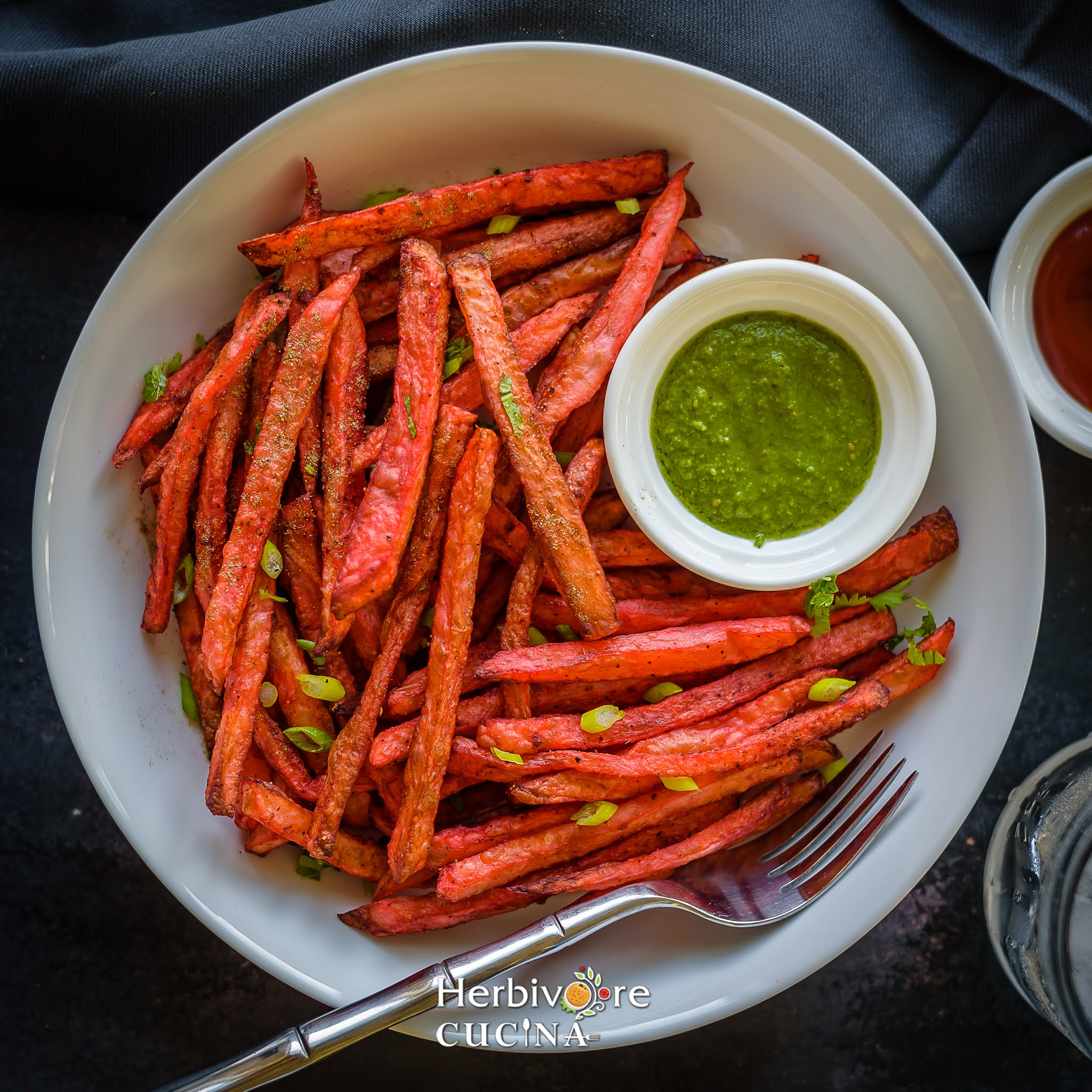 A white plate with a stack of tandoori fries and a bowl of cilantro chutney on a dark background. 