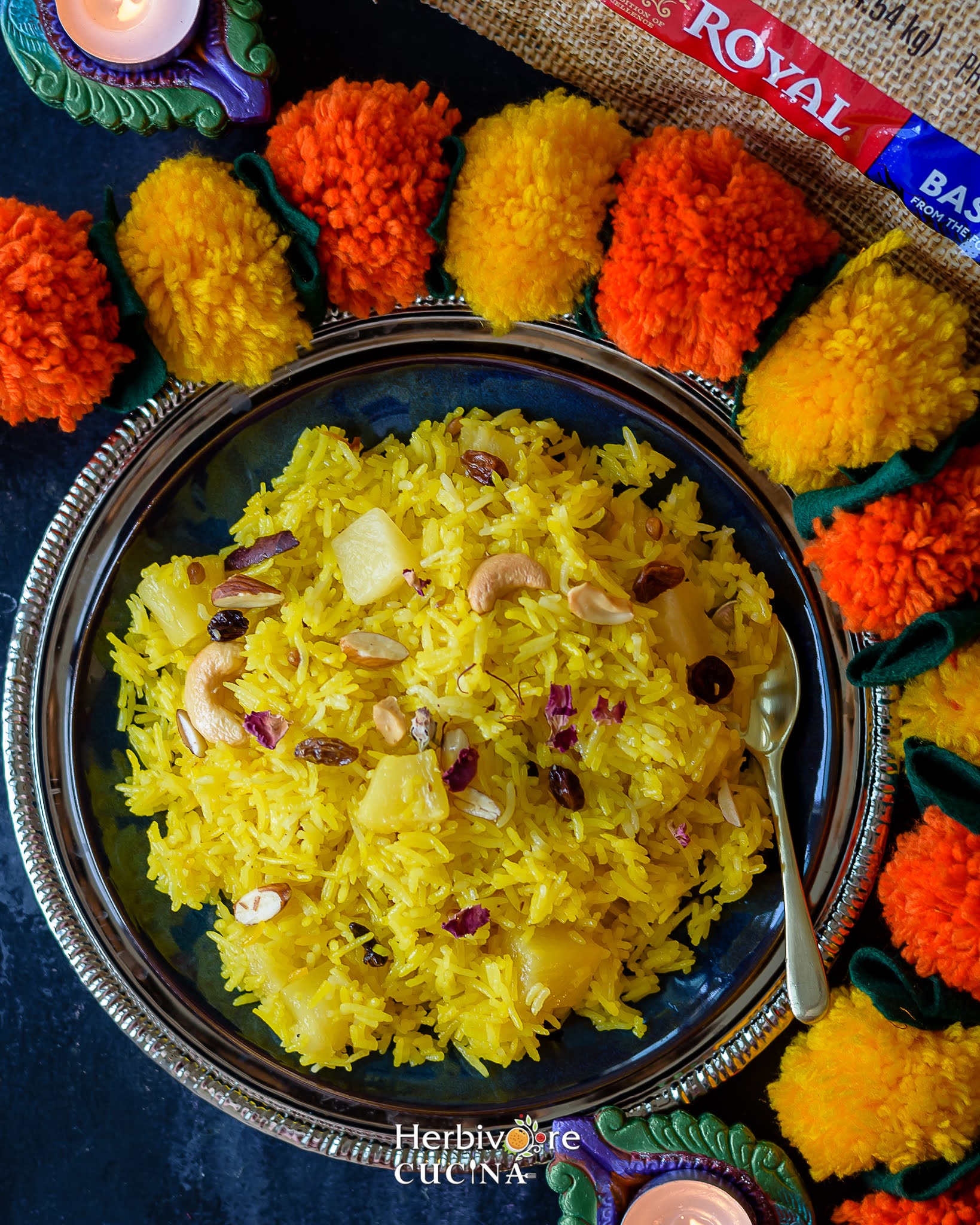 A blue bowl with pineapple zarda rice with flowers around it and a diya by the side on a black board.
