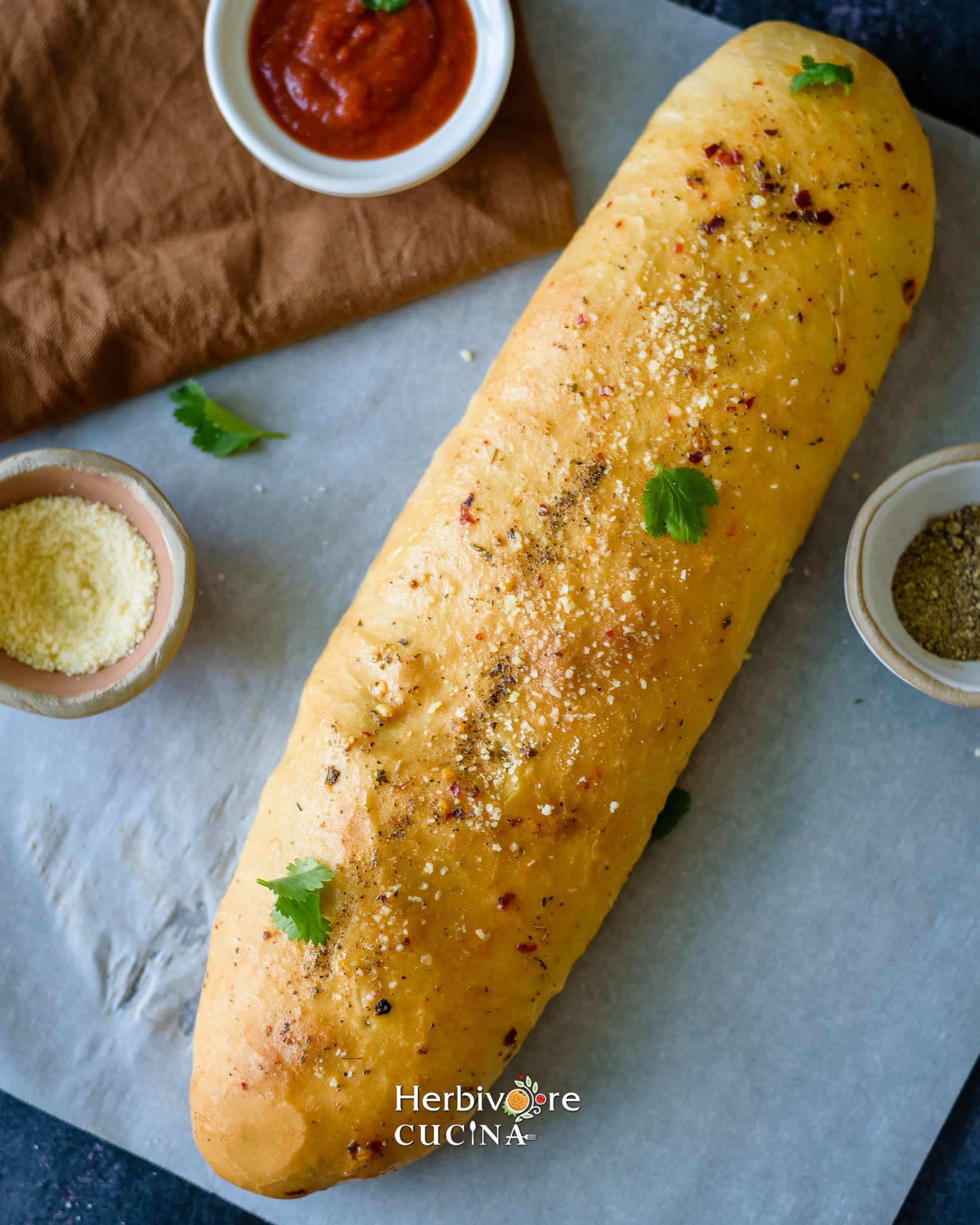 A large log of baked Stromboli with some oregano, chili flakes and cheese around it on a white baking paper. 