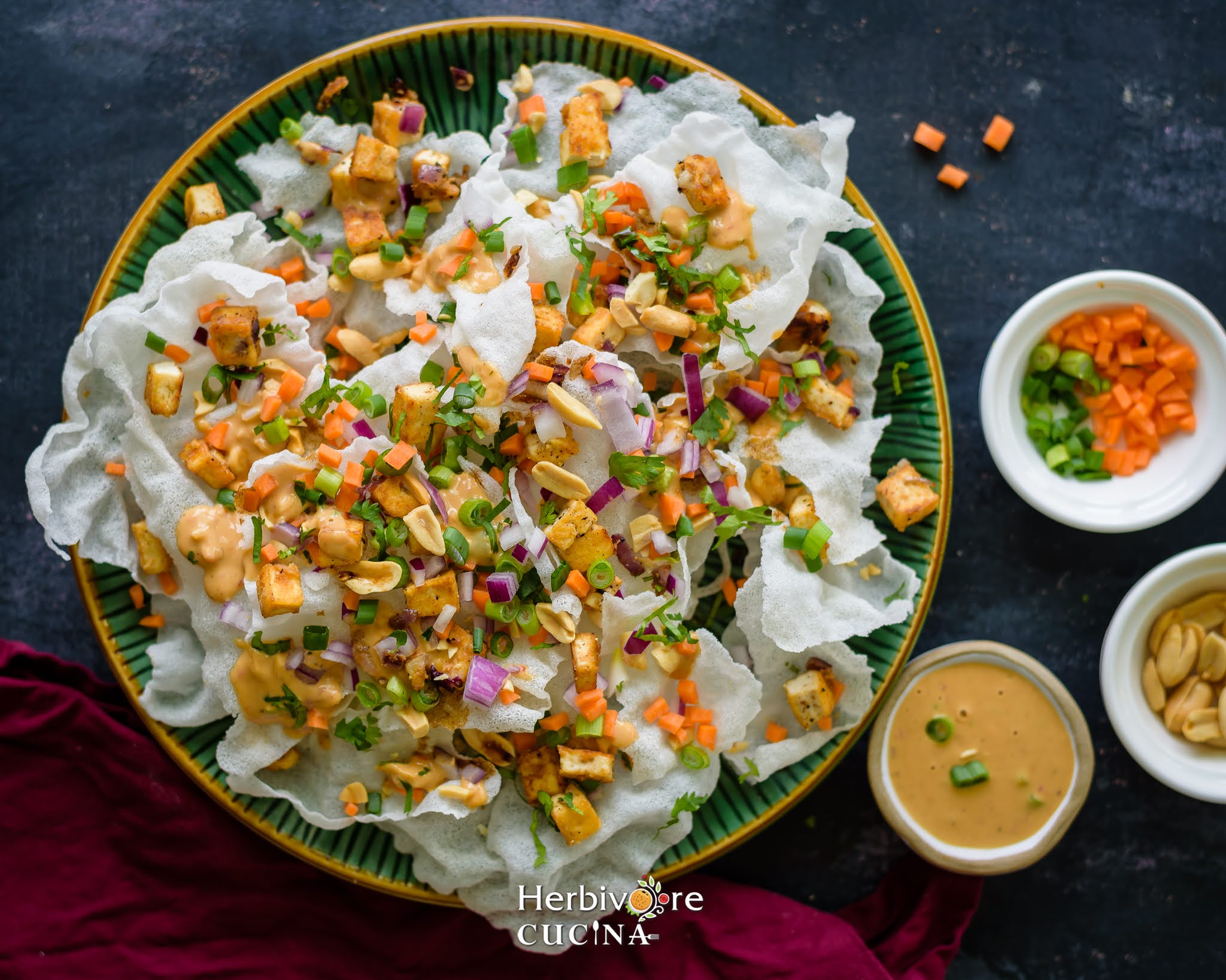 Top view of rice paper crisps in a green plate; topped with tofu, vegetables, peanuts and a peanut based sauce with toppings on the side. 